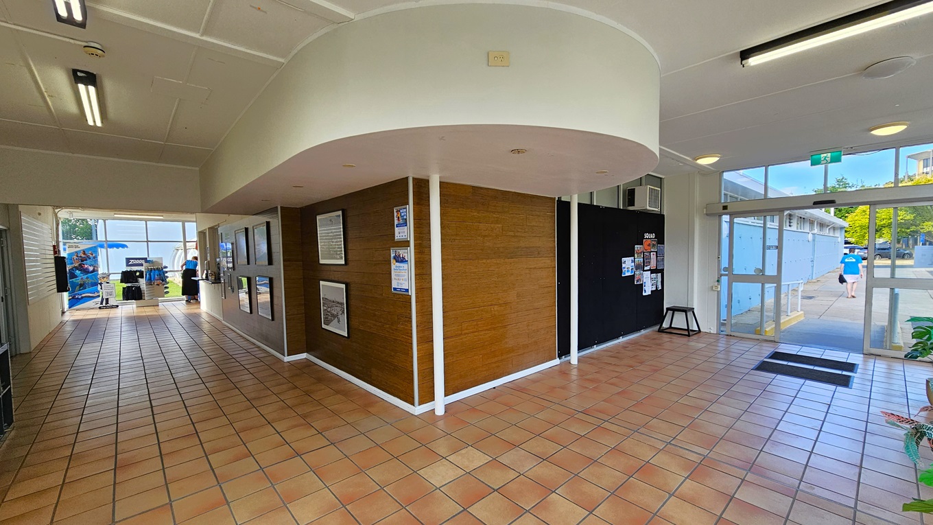 The L-shaped corridor features brown tiles. On the right is the entrance with a glass door and non-slip mats. A blackboard displays posters, and ornamental plants are arranged on the right. The middle wall, with a timber design, showcases large framed posters. At the corridor's end are displayed retail items. The ceiling is white with fluorescent lights. The L-shaped corridor features brown tiles. On the right is the entrance with a glass door and non-slip mats. A blackboard displays posters, and ornamental plants are arranged on the right. The middle wall, with a timber design, showcases large framed posters. At the corridor's end are displayed retail items. The ceiling is white with fluorescent lights.