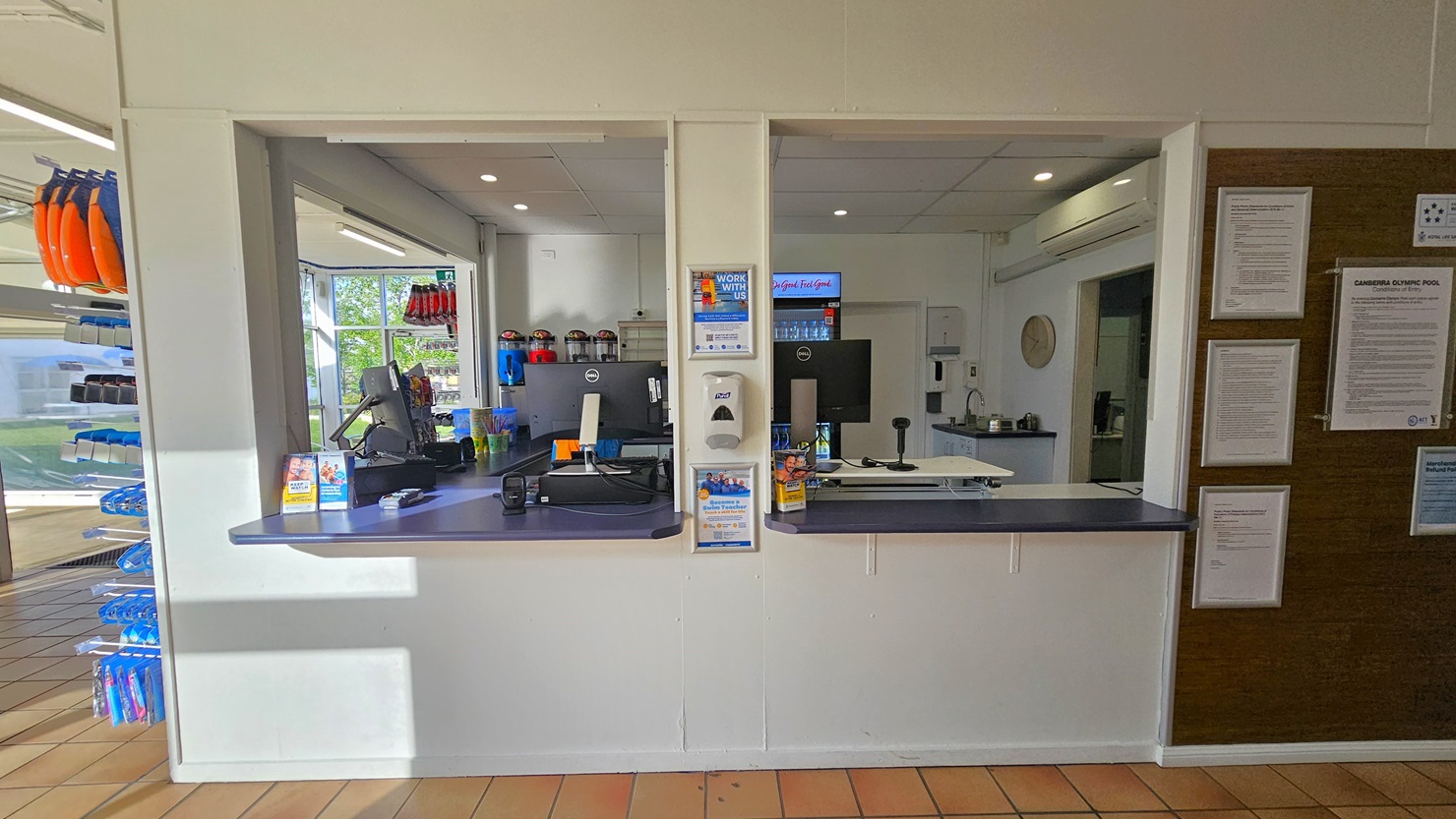 The reception features three large windows. The desk is equipped with computers and swiping machines, with a fridge displaying cold drinks behind. On the right, a brown board displays framed posters, and on the left, a rack showcases goggles and other swimming accessories. The walls and ceiling are white, and the floor has brown tiles. The reception features three large windows. The desk is equipped with computers and swiping machines, with a fridge displaying cold drinks behind. On the right, a brown board displays framed posters, and on the left, a rack showcases goggles and other swimming accessories. The walls and ceiling are white, and the floor has brown tiles.