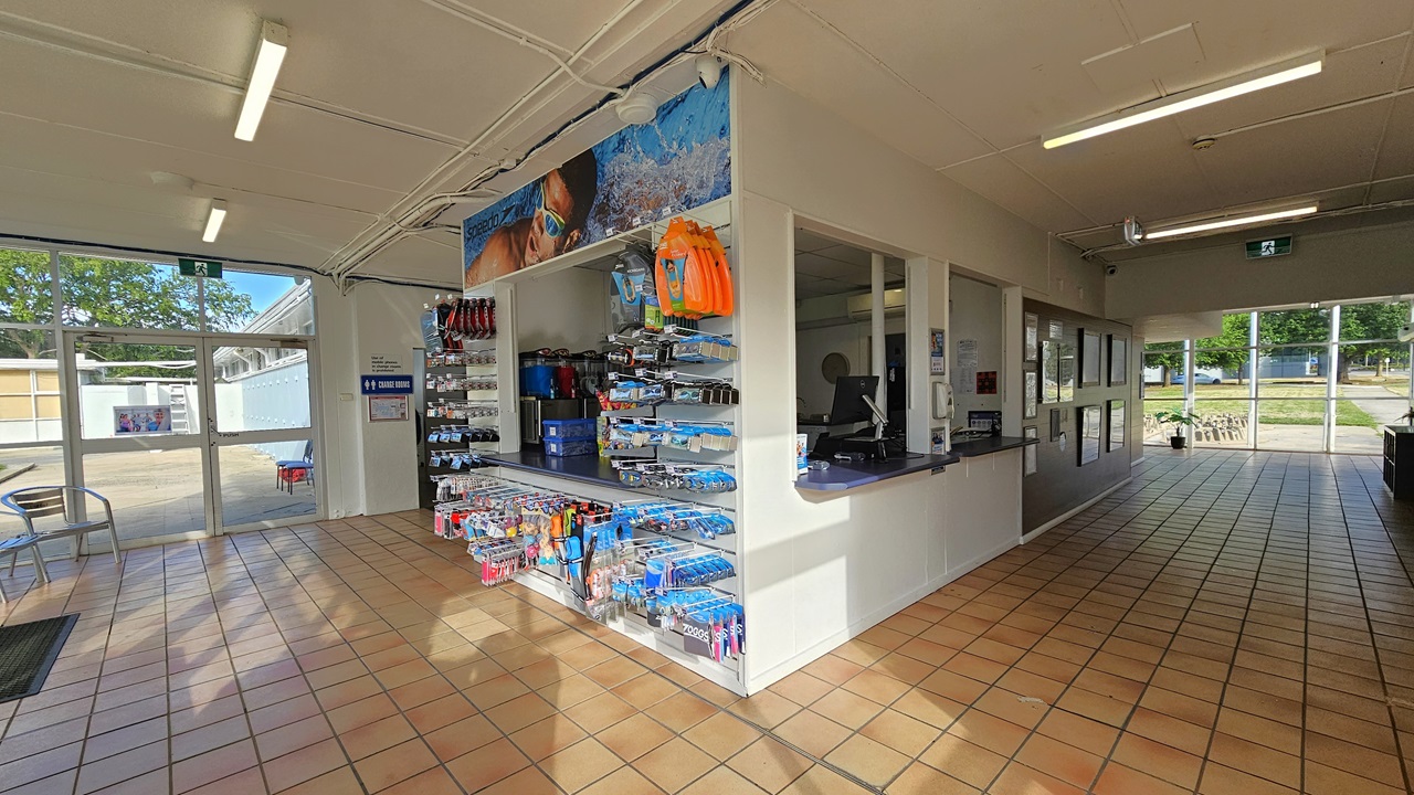 The reception has three large windows. The desk is equipped with computers, swiping machines, and a fridge displaying cold drinks. On the right, a brown board displays framed posters, while the left features a rack showcasing goggles and swimming accessories. Movable chairs and an anti-slip mat are positioned on the right. The walls and ceiling are white, and the floor has brown tiles. The reception has three large windows. The desk is equipped with computers, swiping machines, and a fridge displaying cold drinks. On the right, a brown board displays framed posters, while the left features a rack showcasing goggles and swimming accessories. Movable chairs and an anti-slip mat are positioned on the right. The walls and ceiling are white, and the floor has brown tiles.