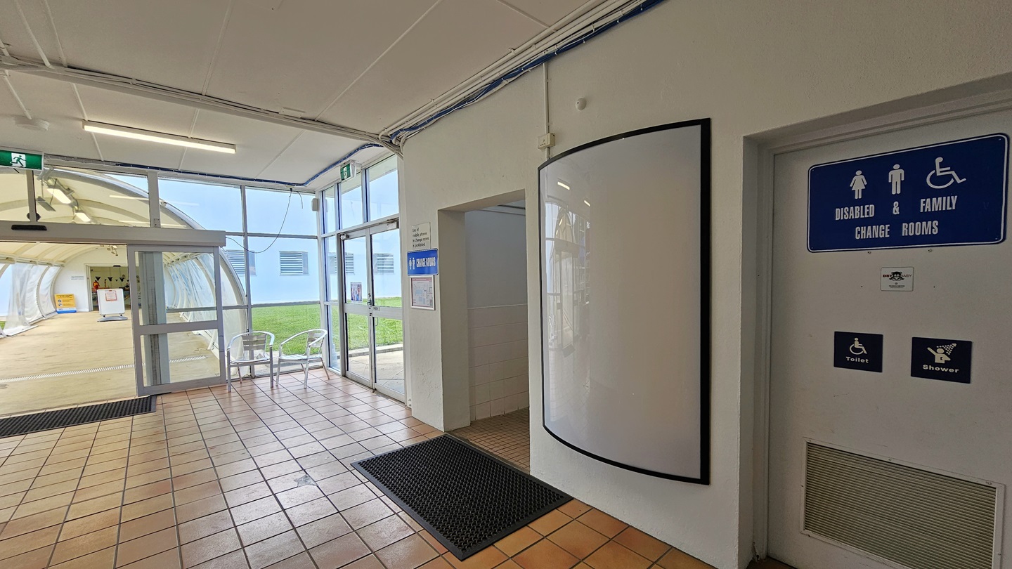 To the right are accessible bathrooms with signage and black rubber mats on the doorsteps. To the left, an automatic glass door leads to a bridge with a dome-shaped roof, featuring two movable chairs in the corner. Another glass door is to the right. The floor has brown tiles, and the ceiling and walls are white. To the right are accessible bathrooms with signage and black rubber mats on the doorsteps. To the left, an automatic glass door leads to a bridge with a dome-shaped roof, featuring two movable chairs in the corner. Another glass door is to the right. The floor has brown tiles, and the ceiling and walls are white.