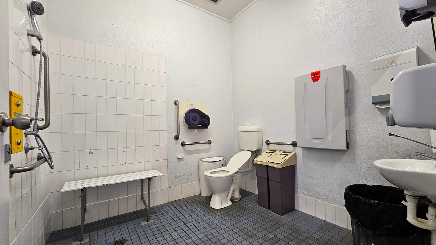 Inside the accessible bathroom, the toilet bowl equipped with handrails is positioned in the corner, accompanied by two rubbish bins. A foldable change table and a tissue dispenser are mounted on the wall. To the left, there is a shower, and adjacent to it is a bench chair. To the right, a wash basin, a liquid soap dispenser, a hand dryer, and a tissue dispenser are mounted on the wall. The walls and ceiling are all white, and the floor has grey tiles. Inside the accessible bathroom, the toilet bowl equipped with handrails is positioned in the corner, accompanied by two rubbish bins. A foldable change table and a tissue dispenser are mounted on the wall. To the left, there is a shower, and adjacent to it is a bench chair. To the right, a wash basin, a liquid soap dispenser, a hand dryer, and a tissue dispenser are mounted on the wall. The walls and ceiling are all white, and the floor has grey tiles.