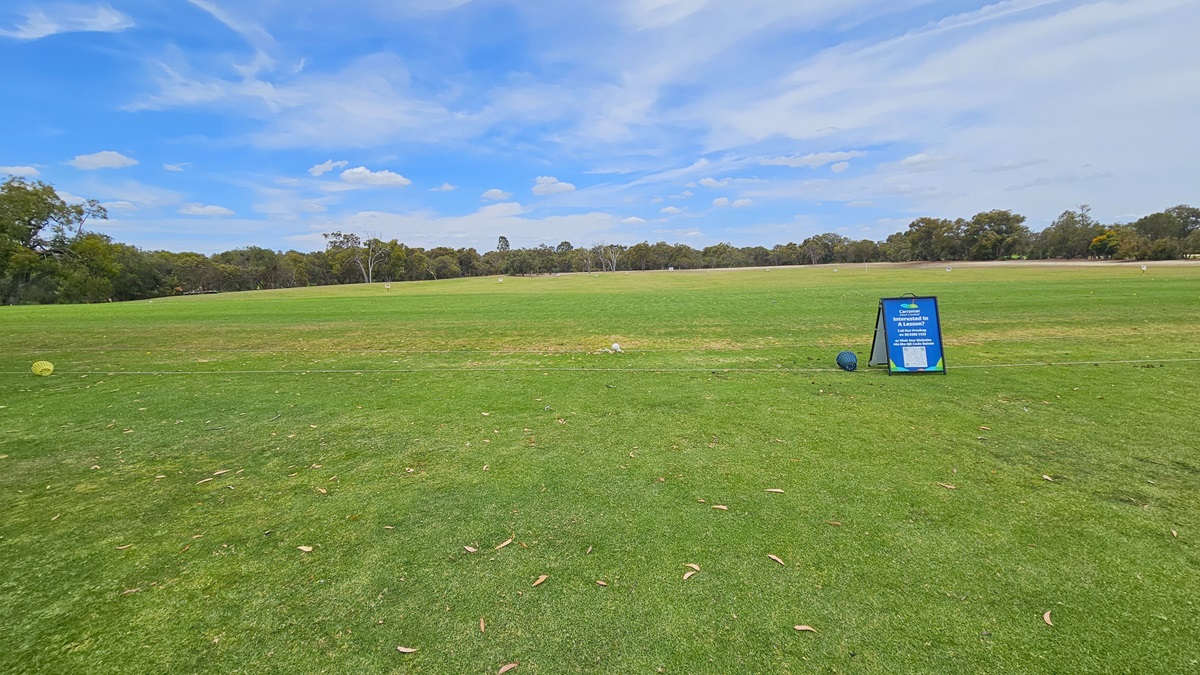 A concrete path directs towards the green. On the right, there is an elevated grassy area with a green sign reading "10th Tee." A golfer is visible near the freestanding banner. On the left, there is manicured grass. Further ahead, more trees are present. A concrete path directs towards the green. On the right, there is an elevated grassy area with a green sign reading "10th Tee." A golfer is visible near the freestanding banner. On the left, there is manicured grass. Further ahead, more trees are present.