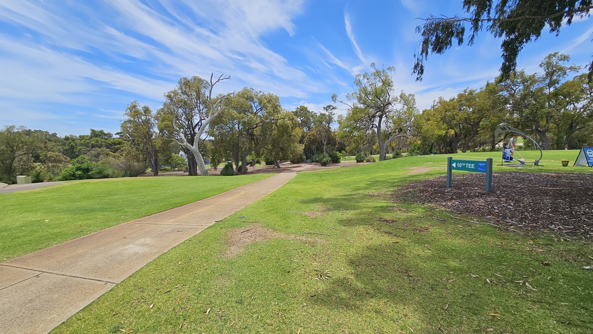 A concrete path directs towards the green. On the right, there is an elevated grassy area with a green sign reading "10th Tee." A golfer is visible near the freestanding banner. On the left, there is manicured grass. Further ahead, more trees are present. A concrete path directs towards the green. On the right, there is an elevated grassy area with a green sign reading "10th Tee." A golfer is visible near the freestanding banner. On the left, there is manicured grass. Further ahead, more trees are present.