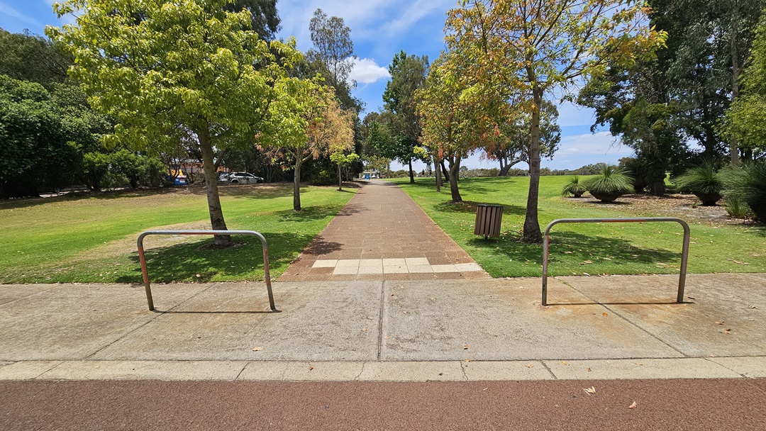 The car park is on a sloping hillside with a pink concrete floor. Three cars of various models and colors are parked under gum trees. A curving concrete path on the right ascends to an elevated area, bordered by tall trees. An accessible parking space, marked with a blue wheelchair symbol, is also visible. The car park is on a sloping hillside with a pink concrete floor. Three cars of various models and colors are parked under gum trees. A curving concrete path on the right ascends to an elevated area, bordered by tall trees. An accessible parking space, marked with a blue wheelchair symbol, is also visible.