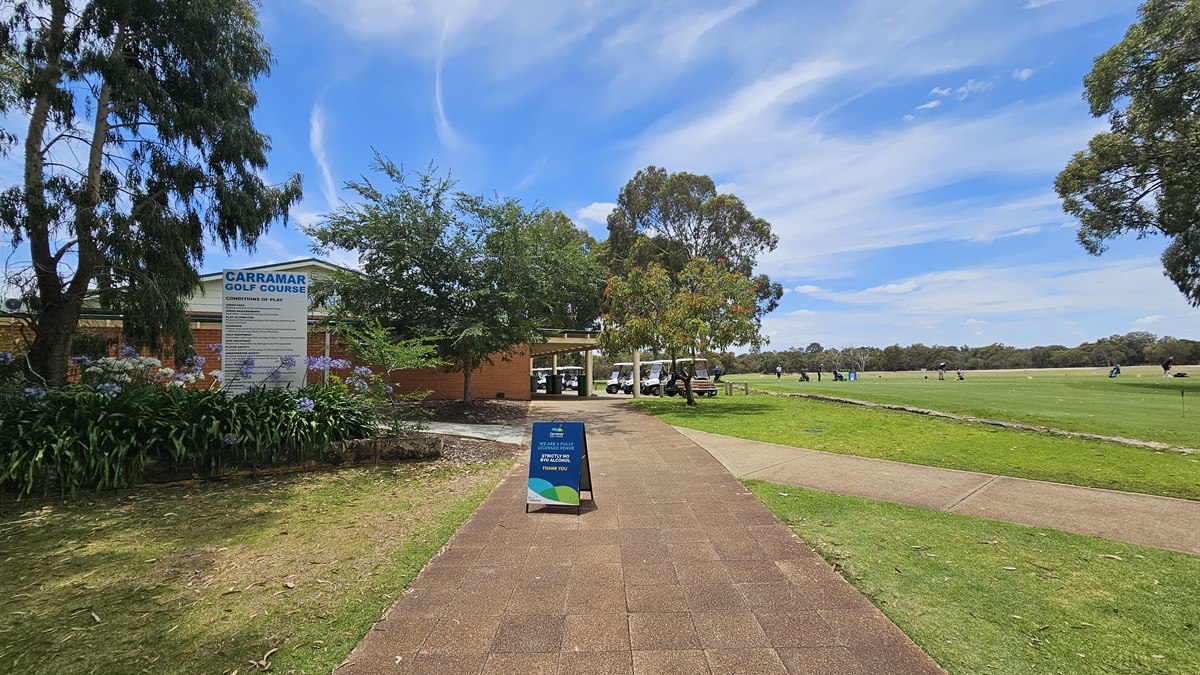On the left stands a red brick building with a veranda. A large banner displaying facility information is positioned on the left. Flowering plants and tall trees border the left side. A blue free standing banner is placed in the middle of the brick pathway. To its right, a path descends to the green, dominating the spacious area. Golf carts are parked near the veranda. On the left stands a red brick building with a veranda. A large banner displaying facility information is positioned on the left. Flowering plants and tall trees border the left side. A blue free standing banner is placed in the middle of the brick pathway. To its right, a path descends to the green, dominating the spacious area. Golf carts are parked near the veranda.