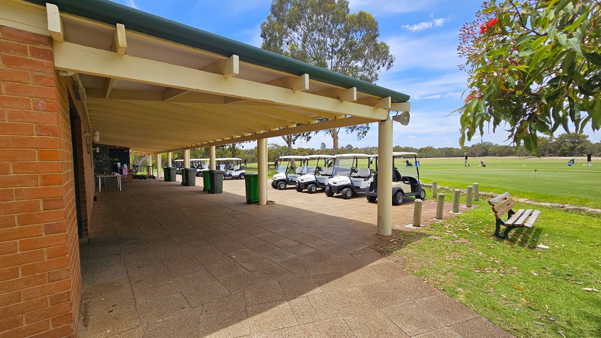 The veranda, constructed with corrugated roofing and exposed wooden beams, has adjacent parked golf carts. Large green rubbish bins, including a white table, are lined in front. The building's facade is made of red bricks. To the right, bench chairs are positioned near concrete bollards. The area on the right features a spacious green where a few golf players are visible. The veranda, constructed with corrugated roofing and exposed wooden beams, has adjacent parked golf carts. Large green rubbish bins, including a white table, are lined in front. The building's facade is made of red bricks. To the right, bench chairs are positioned near concrete bollards. The area on the right features a spacious green where a few golf players are visible.