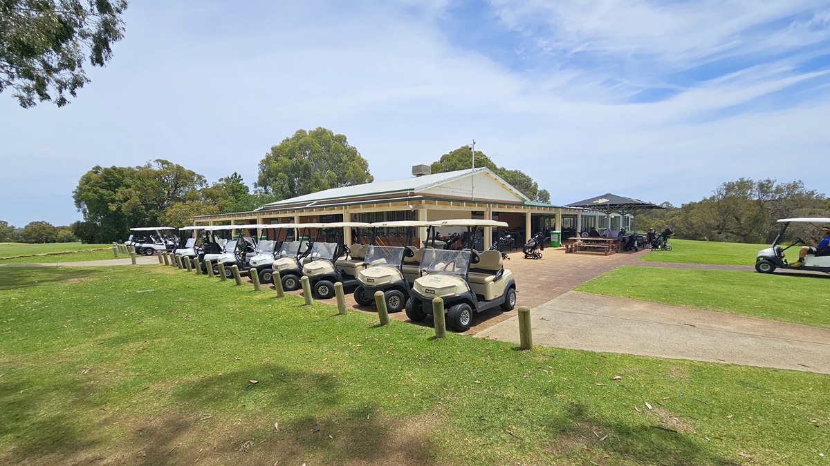 Golf carts are arranged in a row near the sloping grassy area, bordered by concrete bollards. Behind them, a low-rise building with a veranda is equipped with picnic tables under umbrella shades. On the right, a golf cart is parked. The surroundings feature green grass and tall trees. Golf carts are arranged in a row near the sloping grassy area, bordered by concrete bollards. Behind them, a low-rise building with a veranda is equipped with picnic tables under umbrella shades. On the right, a golf cart is parked. The surroundings feature green grass and tall trees.