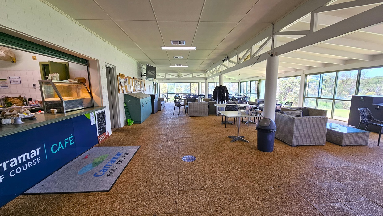 On the left, the cafe counter exhibits the facility's signage with a food rack. Adjacent is a corkboard for reminders, and cleaning accessories are underneath. To the right, there are cane-style couches and movable chairs and tables. A black rubbish bin is positioned near the middle post. The wall is made of white bricks, the ceiling is white, and the floor has brown tiles, featuring a large floor mat with the facility's name. On the left, the cafe counter exhibits the facility's signage with a food rack. Adjacent is a corkboard for reminders, and cleaning accessories are underneath. To the right, there are cane-style couches and movable chairs and tables. A black rubbish bin is positioned near the middle post. The wall is made of white bricks, the ceiling is white, and the floor has brown tiles, featuring a large floor mat with the facility's name.