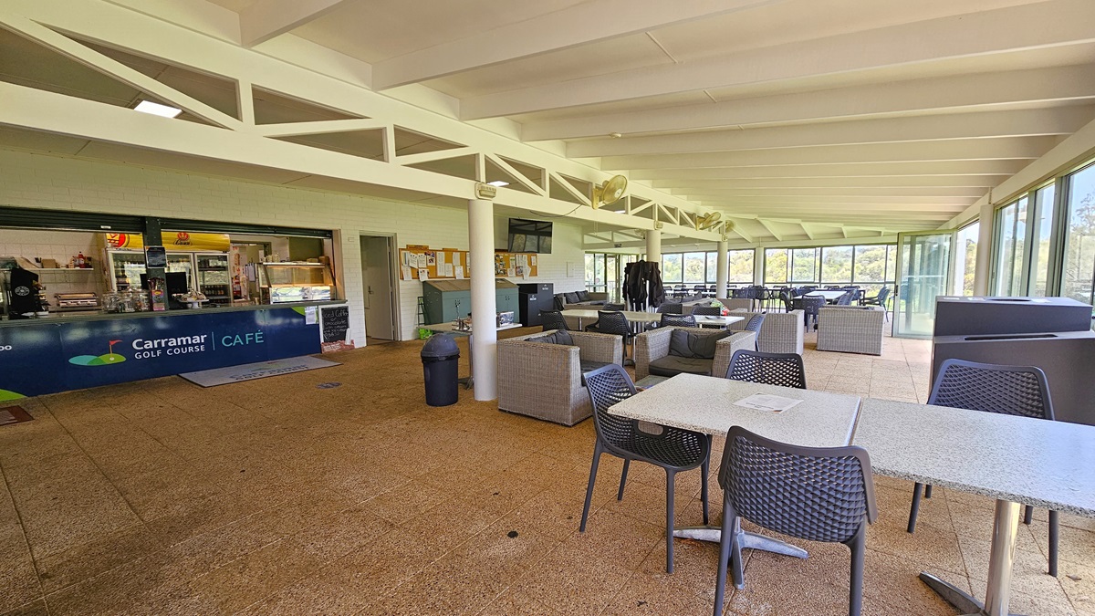 On the left, the cafe counter exhibits the facility's signage with a food rack. Adjacent is a corkboard for reminders, and cleaning accessories are underneath. To the right, there are cane-style couches and movable chairs and tables. A black rubbish bin is positioned near the middle post. The wall is made of white bricks, the ceiling is white, and the floor has brown tiles, featuring a large floor mat with the facility's name. On the left, the cafe counter exhibits the facility's signage with a food rack. Adjacent is a corkboard for reminders, and cleaning accessories are underneath. To the right, there are cane-style couches and movable chairs and tables. A black rubbish bin is positioned near the middle post. The wall is made of white bricks, the ceiling is white, and the floor has brown tiles, featuring a large floor mat with the facility's name.