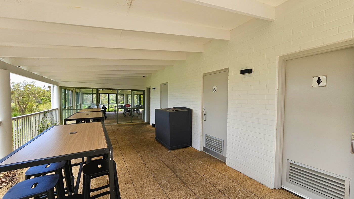 Movable chairs and tables are arranged on the left, secured by a long metal railing. To the right, three grey doors each feature gender access signage. A black rubbish bin is positioned against the white brick wall. The ceiling is white with exposed wooden beams, and the floor is covered with brown tiles. On the far end, a glass door leads to additional seating. Movable chairs and tables are arranged on the left, secured by a long metal railing. To the right, three grey doors each feature gender access signage. A black rubbish bin is positioned against the white brick wall. The ceiling is white with exposed wooden beams, and the floor is covered with brown tiles. On the far end, a glass door leads to additional seating.
