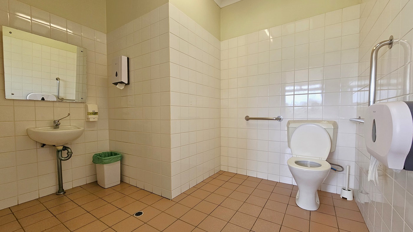 Inside the bathroom, to the left, there is a wash basin with a wall-mounted mirror. A tissue dispenser and a soap dispenser are affixed to the wall, with a rubbish bin underneath. To the right, there is a toilet bowl equipped with handrails. A tissue roll dispenser is also mounted on the right wall. The walls have white tiles, and the floor is made of peach tiles. Inside the bathroom, to the left, there is a wash basin with a wall-mounted mirror. A tissue dispenser and a soap dispenser are affixed to the wall, with a rubbish bin underneath. To the right, there is a toilet bowl equipped with handrails. A tissue roll dispenser is also mounted on the right wall. The walls have white tiles, and the floor is made of peach tiles.