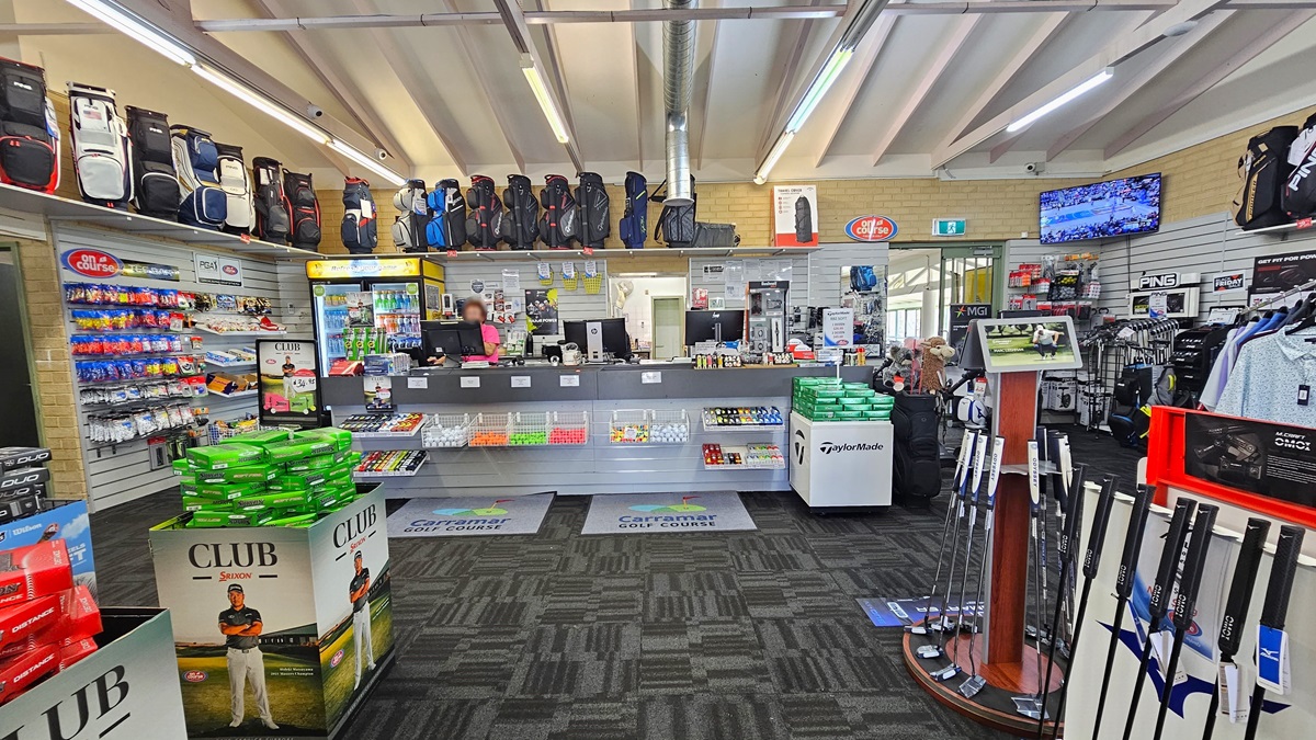 The reception counter, positioned centrally, features three computers and retail items on the desk. A fridge with cold drinks is situated behind the counter. In the upper right corner, a flat-screen TV is mounted. Golf balls and bags are displayed on the left, while golf clubs and polo shirts are arranged on the right, along with additional bags on a display rack. The ceiling is white with exposed metal beams and fluorescent lights, and the floor is covered with grey carpet. The reception counter, positioned centrally, features three computers and retail items on the desk. A fridge with cold drinks is situated behind the counter. In the upper right corner, a flat-screen TV is mounted. Golf balls and bags are displayed on the left, while golf clubs and polo shirts are arranged on the right, along with additional bags on a display rack. The ceiling is white with exposed metal beams and fluorescent lights, and the floor is covered with grey carpet.