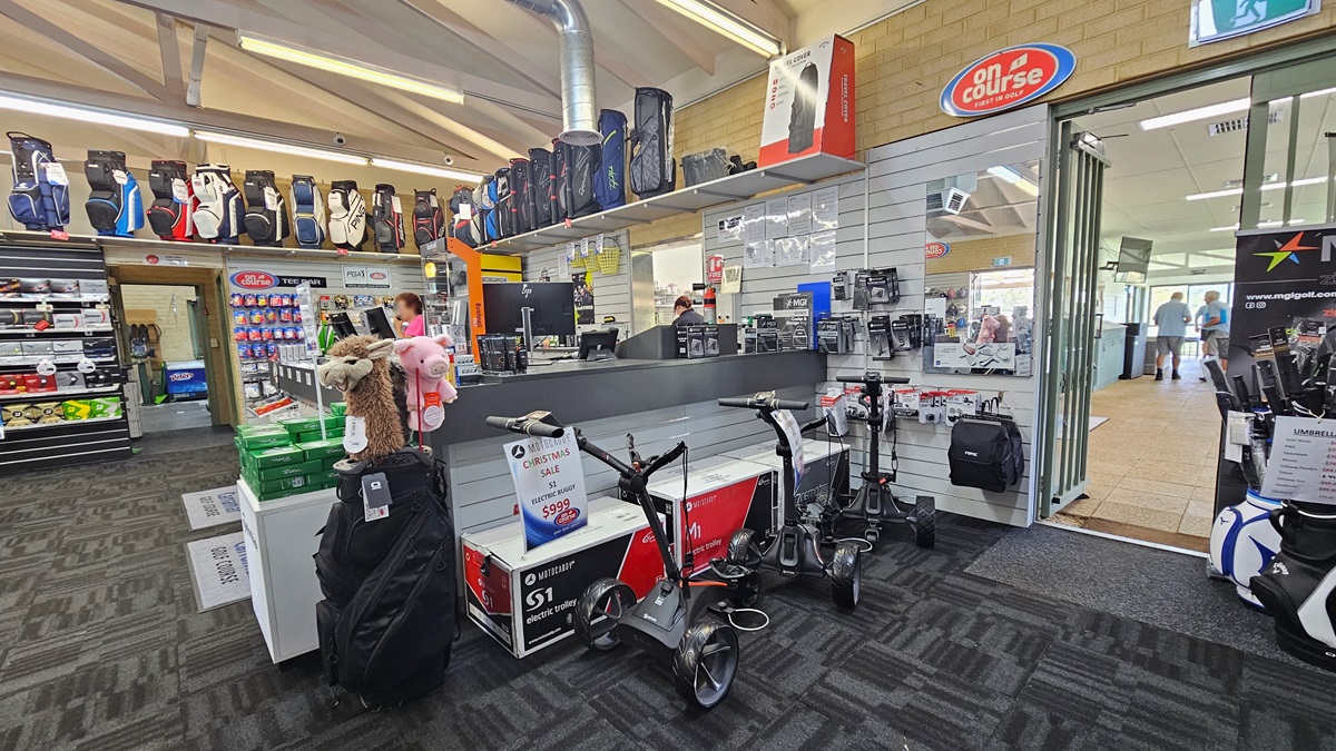 The reception counter, centrally positioned, accommodates three computers and retail items. Golf balls and bags are displayed on the left, while electric trolleys are showcased on the right. A glass door on the right opens outward, leading to an area with cream tiles. The ceiling is white with exposed metal beams and fluorescent lights, and the floor is covered with grey carpet. The reception counter, centrally positioned, accommodates three computers and retail items. Golf balls and bags are displayed on the left, while electric trolleys are showcased on the right. A glass door on the right opens outward, leading to an area with cream tiles. The ceiling is white with exposed metal beams and fluorescent lights, and the floor is covered with grey carpet.