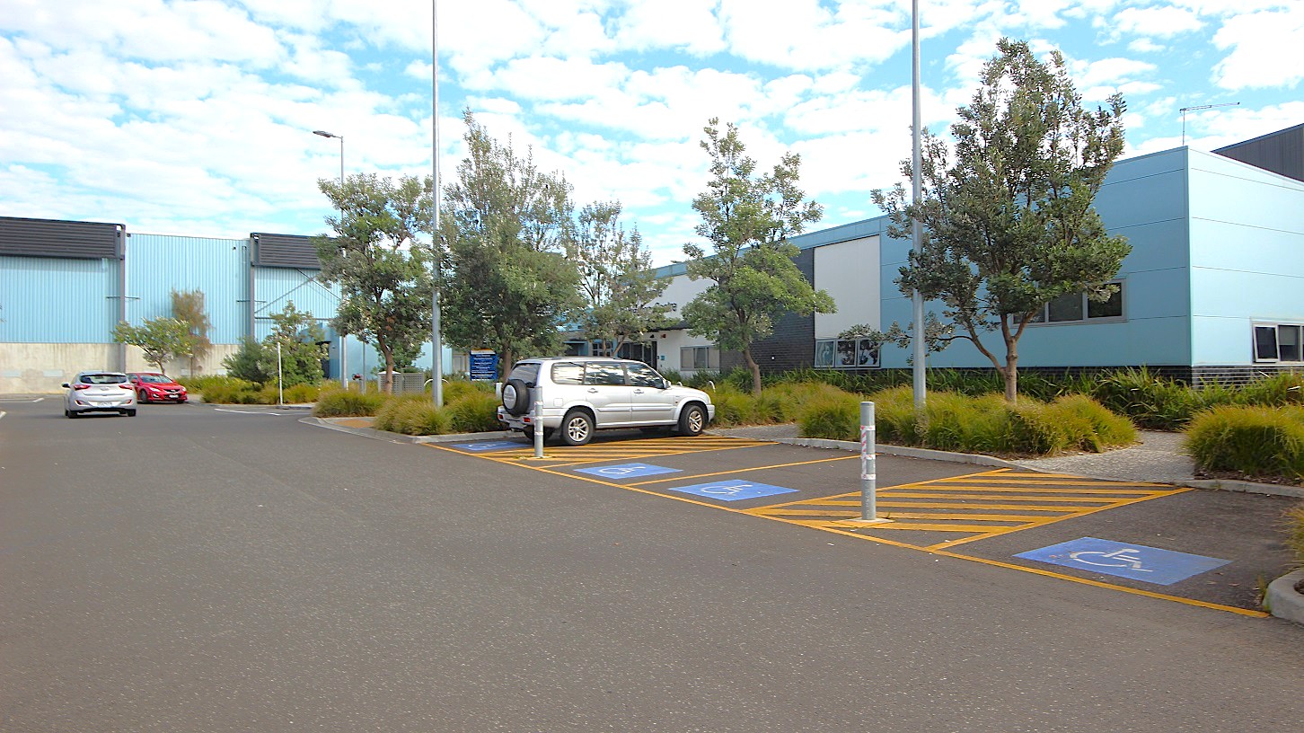 In the foreground, there is a large, paved parking area with multiple spaces marked, including two designated for disabled parking. A white vehicle is parked in one of these spaces. To the left, a grey car is visible further down the parking area. On the right, the building features blue cladding with several windows, surrounded by landscaped areas with small trees and shrubs. Several light poles are scattered throughout the parking lot, providing illumination. In the foreground, there is a large, paved parking area with multiple spaces marked, including two designated for disabled parking. A white vehicle is parked in one of these spaces. To the left, a grey car is visible further down the parking area. On the right, the building features blue cladding with several windows, surrounded by landscaped areas with small trees and shrubs. Several light poles are scattered throughout the parking lot, providing illumination.