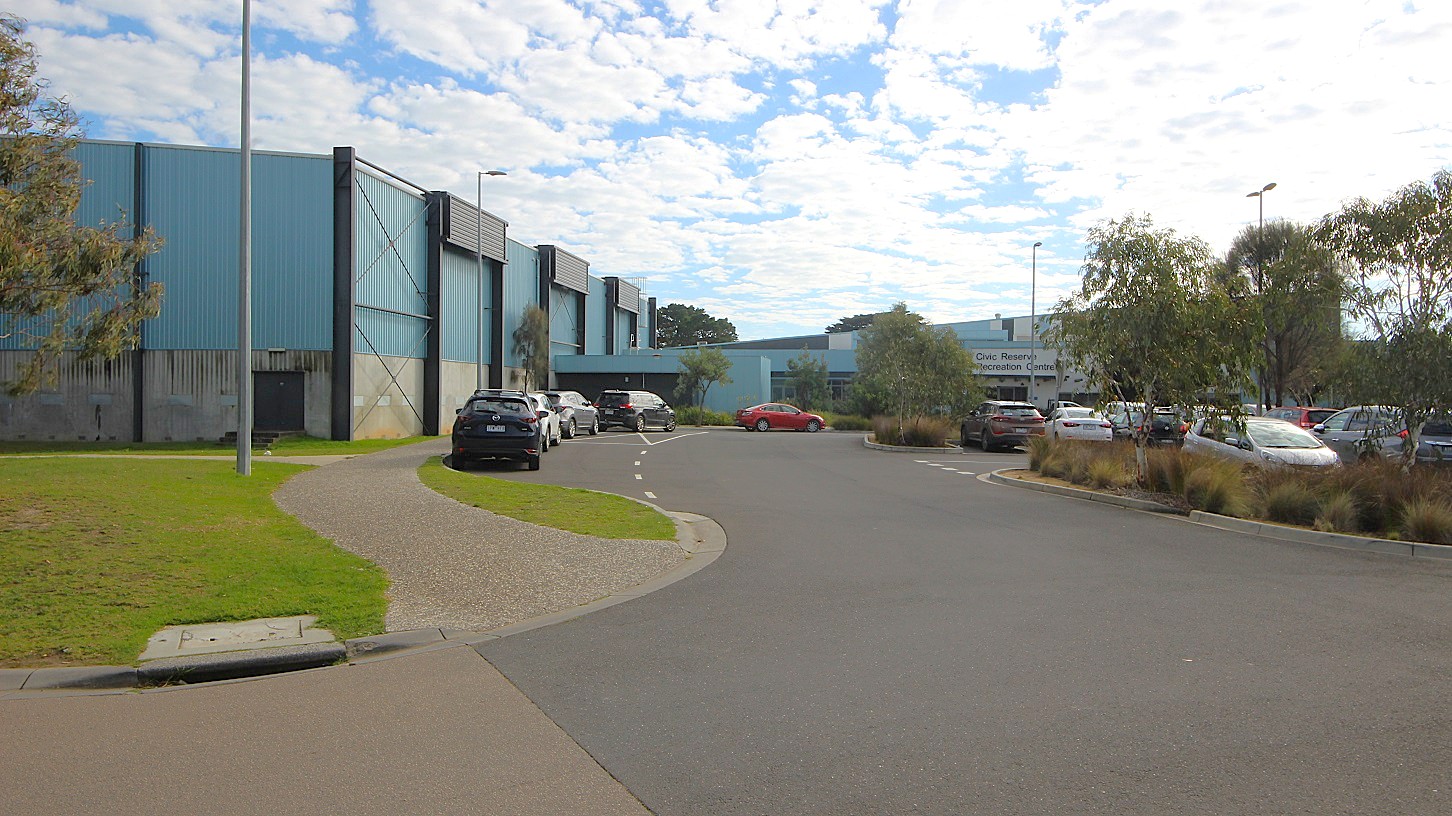 In the foreground, a curved asphalt road leads past landscaped grass areas on the left, bordered by a gravel path. Several vehicles are parked along the right side of the road, with one black car prominently displayed. To the left, the building features a blue exterior with vertical paneling, and there are light poles positioned throughout the area. In the background, the Civic Reserve Recreation Centre is visible, identified by its signage, with more vehicles parked in a lot beyond. In the foreground, a curved asphalt road leads past landscaped grass areas on the left, bordered by a gravel path. Several vehicles are parked along the right side of the road, with one black car prominently displayed. To the left, the building features a blue exterior with vertical paneling, and there are light poles positioned throughout the area. In the background, the Civic Reserve Recreation Centre is visible, identified by its signage, with more vehicles parked in a lot beyond.