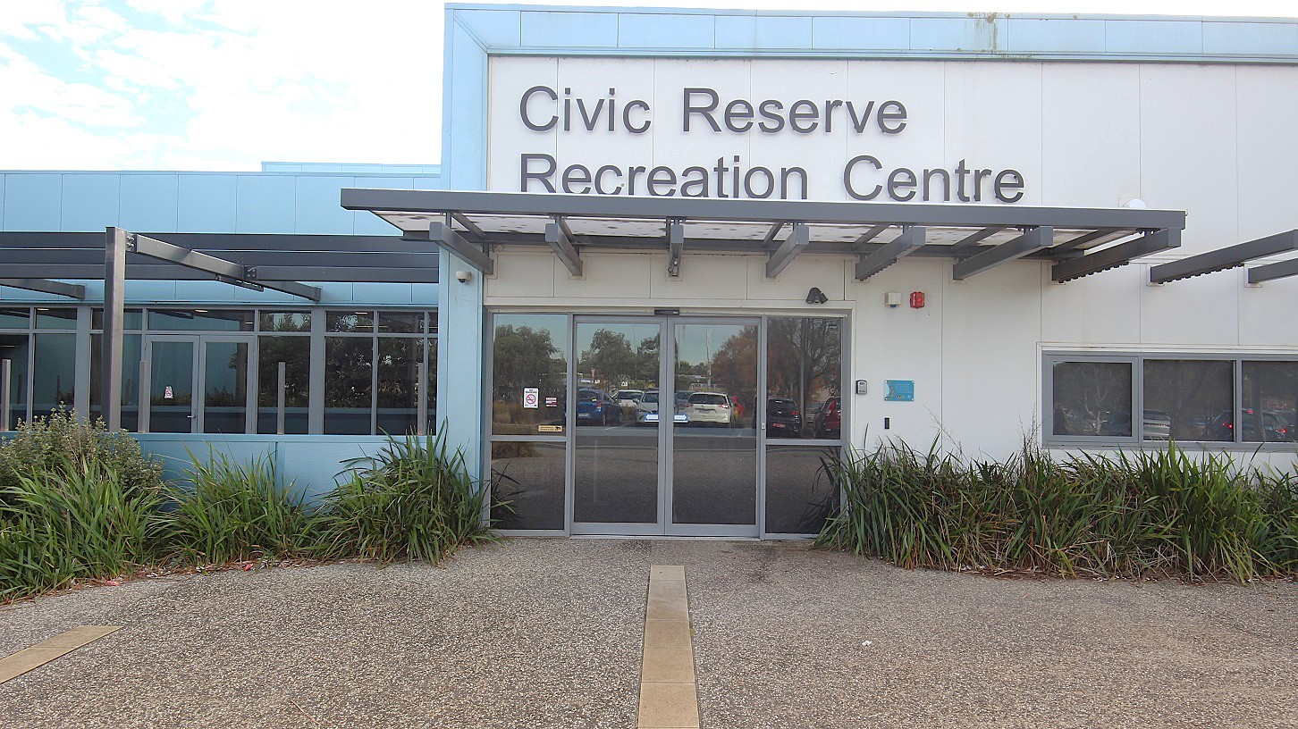 A closer look at the entrance glass doors with light grey painted edges and an overhang made of grey metal with a white membrane and transparent roofing at Civic Reserve Recreation Centre. Near the venue, there is a carpark where the glass reflects the cars parked in the middle. On the left door, there is a no-smoking sign. The name of the venue can be seen at the top. Covering the lower portion of the walls of the venue are plants with parallel venation. A closer look at the entrance glass doors with light grey painted edges and an overhang made of grey metal with a white membrane and transparent roofing at Civic Reserve Recreation Centre. Near the venue, there is a carpark where the glass reflects the cars parked in the middle. On the left door, there is a no-smoking sign. The name of the venue can be seen at the top. Covering the lower portion of the walls of the venue are plants with parallel venation.