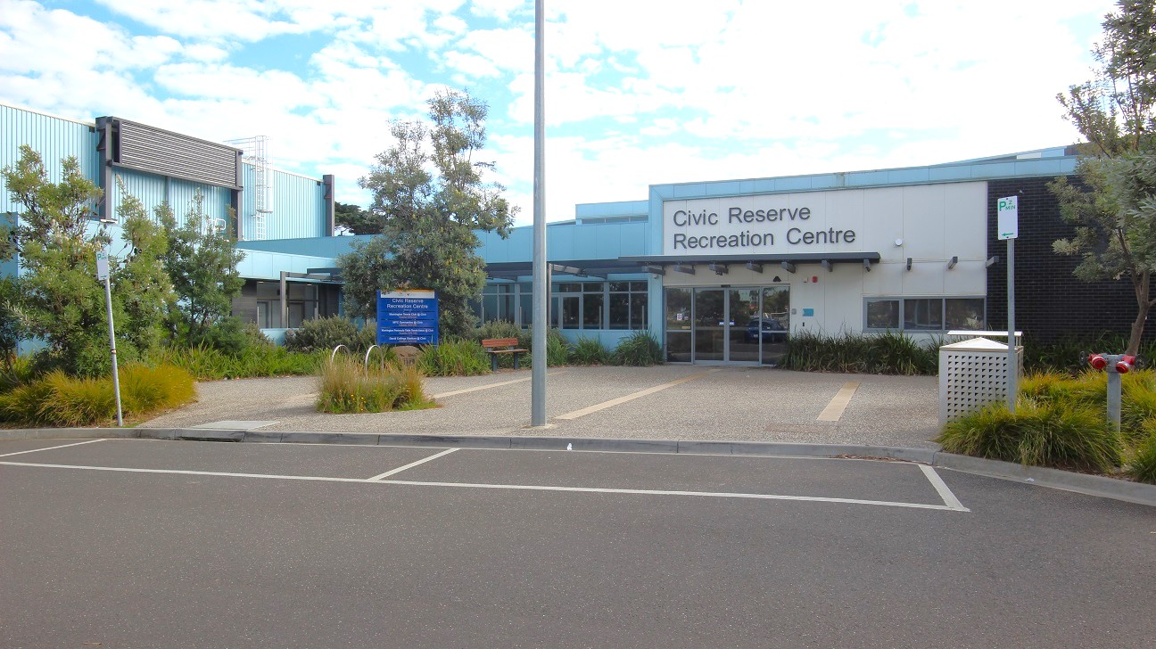 In the foreground, there is a paved area leading to the entrance of the Civic Reserve Recreation Centre, which features large glass windows and a prominent sign. To the left, a gravel path lined with greenery is visible, along with several small shrubs. On the right, there is a decorative planter and a parking sign. The ground is made of light-coloured stones interspersed with grass, providing a neat appearance. In the foreground, there is a paved area leading to the entrance of the Civic Reserve Recreation Centre, which features large glass windows and a prominent sign. To the left, a gravel path lined with greenery is visible, along with several small shrubs. On the right, there is a decorative planter and a parking sign. The ground is made of light-coloured stones interspersed with grass, providing a neat appearance.