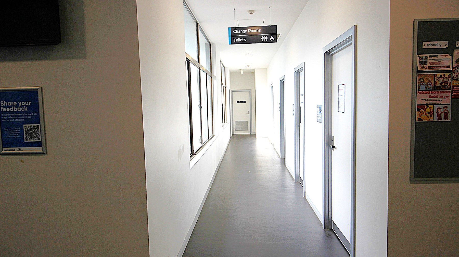 Hallway to the changing rooms and bathrooms at Civic Reserve Recreation Centre. The walls are white, and the floor is grey. On the opposite side of the 4 doors on the right with sky-blue painted edges are glass windows. Hanging on the ceiling is a small black signboard for Change rooms and Toilets. Hallway to the changing rooms and bathrooms at Civic Reserve Recreation Centre. The walls are white, and the floor is grey. On the opposite side of the 4 doors on the right with sky-blue painted edges are glass windows. Hanging on the ceiling is a small black signboard for Change rooms and Toilets.