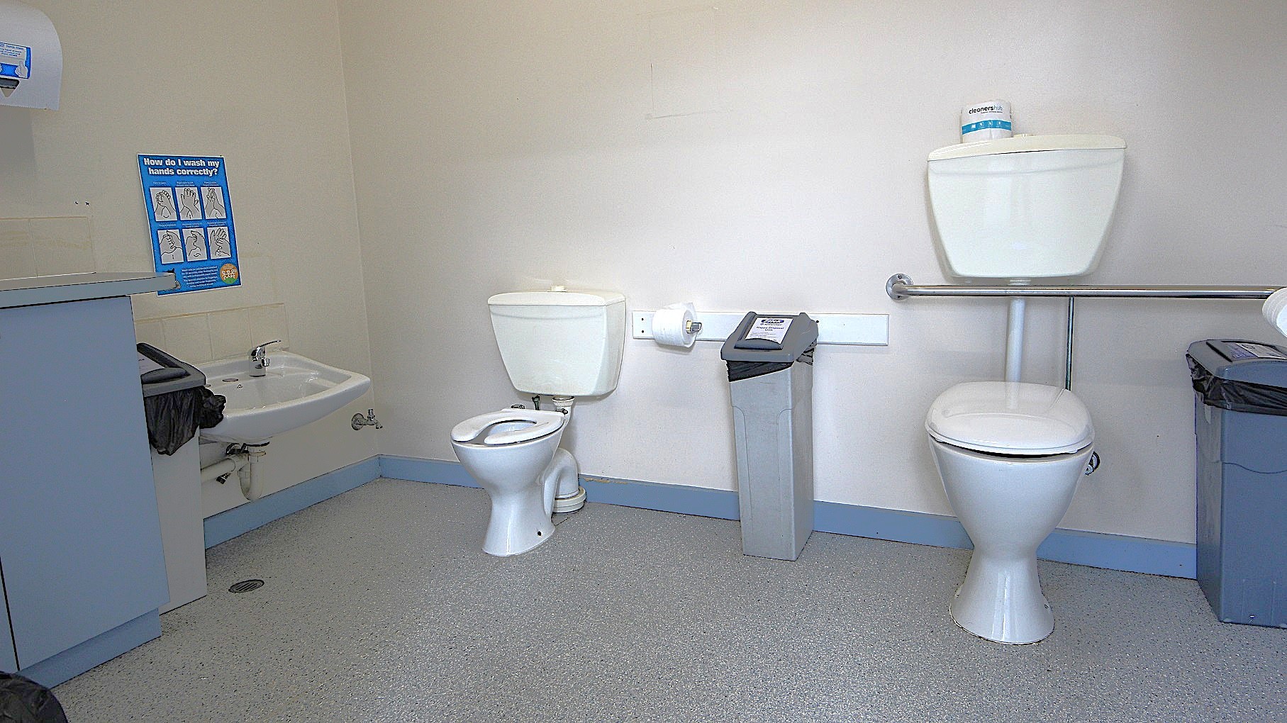 Inside the accessible bathroom at Civic Reserve Recreation Centre. On the left wall is a paper towel dispenser, a grey rubbish bin, a low-set wash basin, and a blue infographic about how to wash hands properly. In the middle are two toilet seats of different heights and sizes (left small; right big). Between them is a sanitary napkin bin and a toilet roll holder with toilet paper. The floor is granite, and the walls are painted white. Inside the accessible bathroom at Civic Reserve Recreation Centre. On the left wall is a paper towel dispenser, a grey rubbish bin, a low-set wash basin, and a blue infographic about how to wash hands properly. In the middle are two toilet seats of different heights and sizes (left small; right big). Between them is a sanitary napkin bin and a toilet roll holder with toilet paper. The floor is granite, and the walls are painted white.