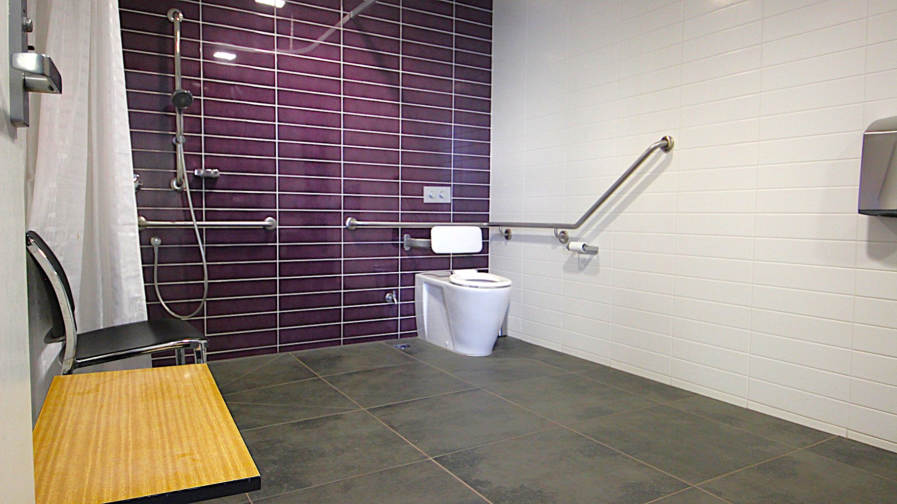 Image inside one of the accessible bathrooms at Civic Reserve Recreation Centre. On the left is a knee-high wooden table and a black chair next to it. There is a white shower curtain, a handheld showerhead, and grab bars situated on the purple-tiled wall. and to the right is a toilet. The bathroom has a grey-tiled floor and a white-painted brick wall. Image inside one of the accessible bathrooms at Civic Reserve Recreation Centre. On the left is a knee-high wooden table and a black chair next to it. There is a white shower curtain, a handheld showerhead, and grab bars situated on the purple-tiled wall. and to the right is a toilet. The bathroom has a grey-tiled floor and a white-painted brick wall.