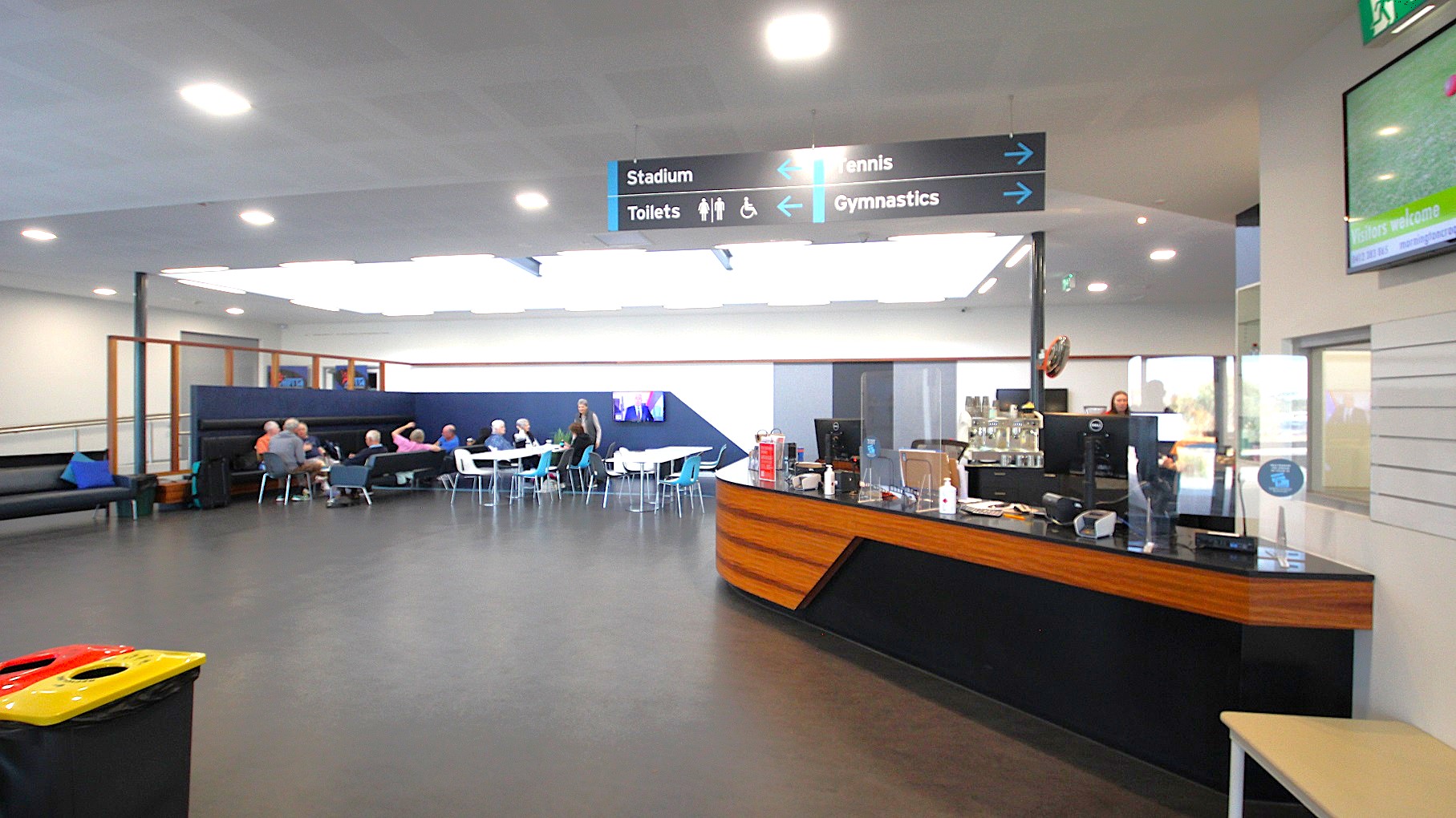 The first section inside the Civic Reserve Recreation Centre is a black and walnut counter of the café and seating area occupied by adults enjoying themselves while chatting. Lighting the seating area is a skylight allowing natural light to filter in addition to fluorescent strips that surround this rectangular skylight. There is black signage indicating the location of the Stadium, Toilets, Tennis, and Gymnasium. The first section inside the Civic Reserve Recreation Centre is a black and walnut counter of the café and seating area occupied by adults enjoying themselves while chatting. Lighting the seating area is a skylight allowing natural light to filter in addition to fluorescent strips that surround this rectangular skylight. There is black signage indicating the location of the Stadium, Toilets, Tennis, and Gymnasium.
