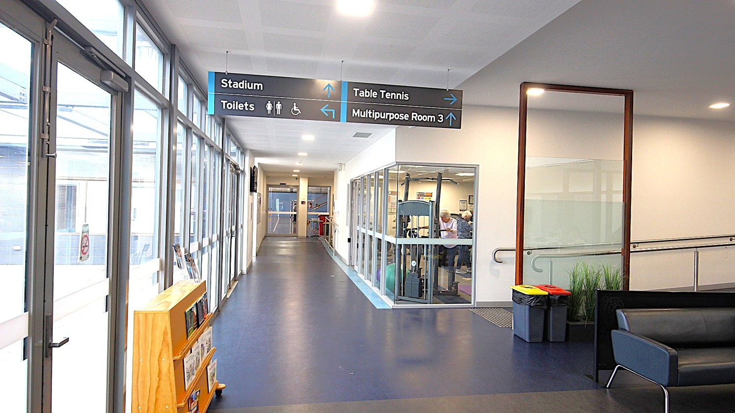 Hallway on the left side of the main entrance doors. The image shows a wooden brown cabinet behind the glass doors on the left, a low black sofa chair, and black rubbish bins with red and yellow lids at the start of the entrance ramp on the right. Looking straight ahead, this hallway has a grey floor, a room to the right filled with adults using exercising equipment, and hand rails on the white wall. Hallway on the left side of the main entrance doors. The image shows a wooden brown cabinet behind the glass doors on the left, a low black sofa chair, and black rubbish bins with red and yellow lids at the start of the entrance ramp on the right. Looking straight ahead, this hallway has a grey floor, a room to the right filled with adults using exercising equipment, and hand rails on the white wall.