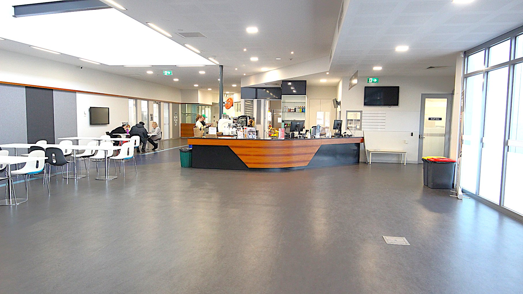 The image shows the side of the black and walnut café counter and its surrounding area. On the right side is a green fire exit sign. Below it is a television mounted on the upper corner of the wall, and next is the “Staff only” white, single, glass door. On the left is a seating area with white chairs and tables that have steel legs and frame. The image shows the side of the black and walnut café counter and its surrounding area. On the right side is a green fire exit sign. Below it is a television mounted on the upper corner of the wall, and next is the “Staff only” white, single, glass door. On the left is a seating area with white chairs and tables that have steel legs and frame.