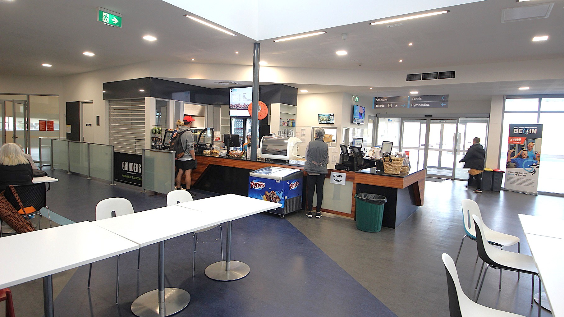 At the café counter is a black cash register, a glass display cabinet with mouth-watering pastries, and small, well-designed flyers with aesthetic colors. There are cold drinks stored in a large fridge, and the menu can be seen on the digital screen mounted on the wall above it. Further to the left is a separate walkway by grey stanchions towards a white door room next to the café. At the café counter is a black cash register, a glass display cabinet with mouth-watering pastries, and small, well-designed flyers with aesthetic colors. There are cold drinks stored in a large fridge, and the menu can be seen on the digital screen mounted on the wall above it. Further to the left is a separate walkway by grey stanchions towards a white door room next to the café.