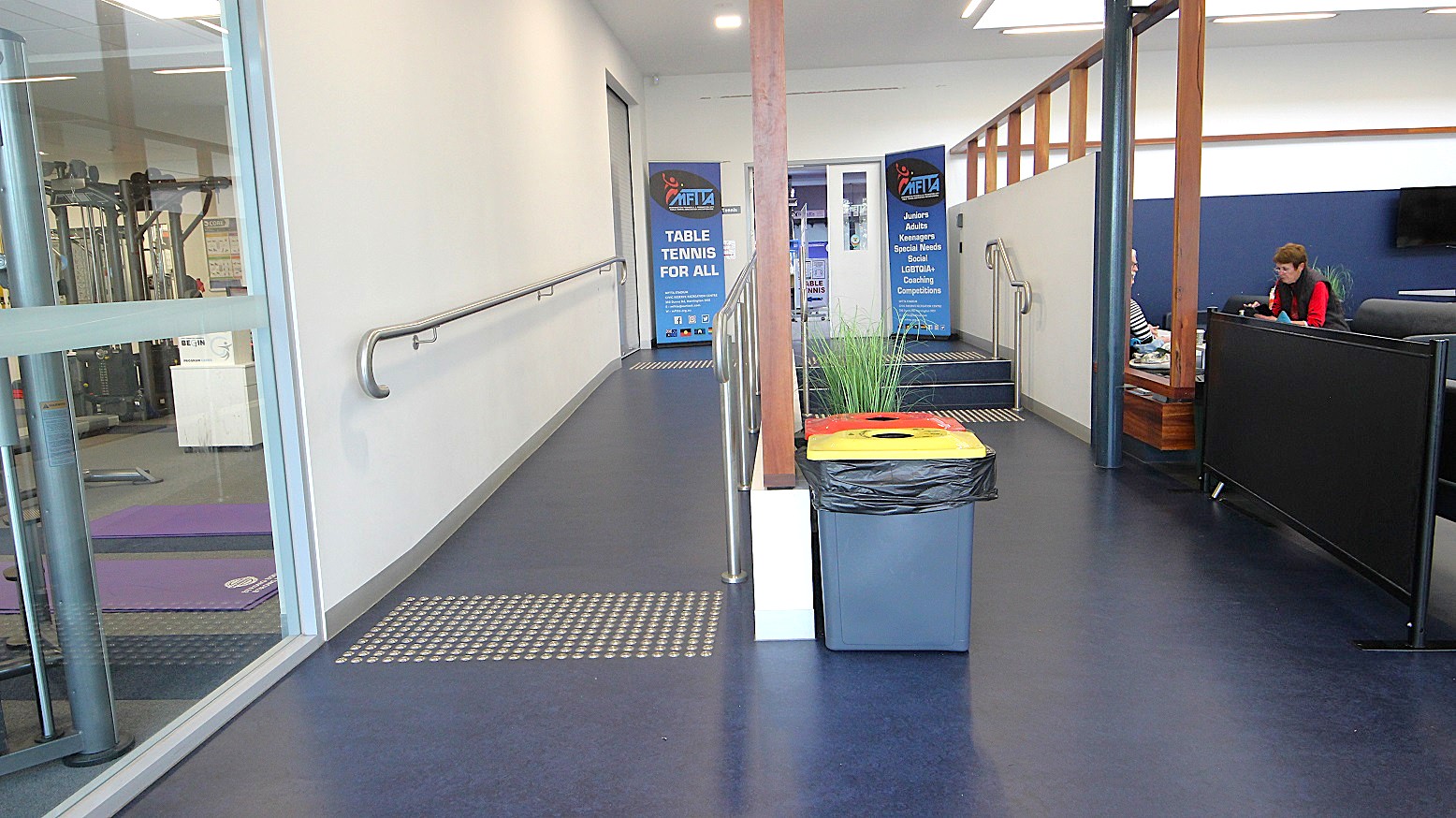 Entry to the table tennis area is via the entrance ramp and a two-step tier stairwell, both of which have handrails to assist individuals with mobility disabilities. The starting points of the ramp and stairwell are designed with tactile ground surface indicators. Potted plants and black rubbish bins with yellow and orange lids are located in front of the stairwell. Entry to the table tennis area is via the entrance ramp and a two-step tier stairwell, both of which have handrails to assist individuals with mobility disabilities. The starting points of the ramp and stairwell are designed with tactile ground surface indicators. Potted plants and black rubbish bins with yellow and orange lids are located in front of the stairwell.