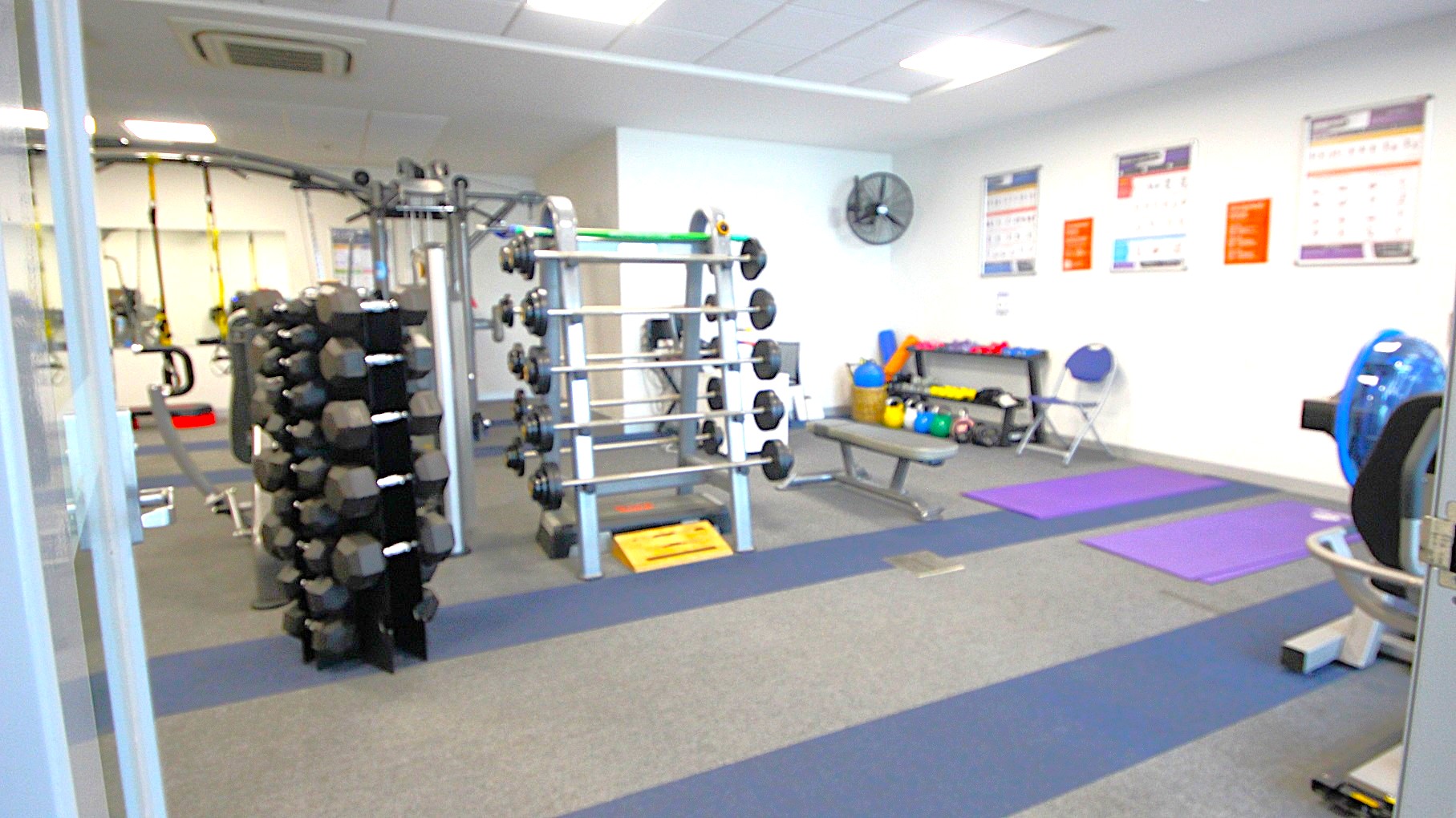 A gym with various equipment. On the left, there is a rack of dumbbells. In the centre, a weightlifting station with multiple barbells. On the right, exercise mats are laid out on the floor, with a rowing machine partially visible. The back wall has posters and a fan, with a bench holding kettlebells and medicine balls. The floor is carpeted in grey and blue stripes, and the ceiling has recessed lighting and an air conditioning unit. A gym with various equipment. On the left, there is a rack of dumbbells. In the centre, a weightlifting station with multiple barbells. On the right, exercise mats are laid out on the floor, with a rowing machine partially visible. The back wall has posters and a fan, with a bench holding kettlebells and medicine balls. The floor is carpeted in grey and blue stripes, and the ceiling has recessed lighting and an air conditioning unit.