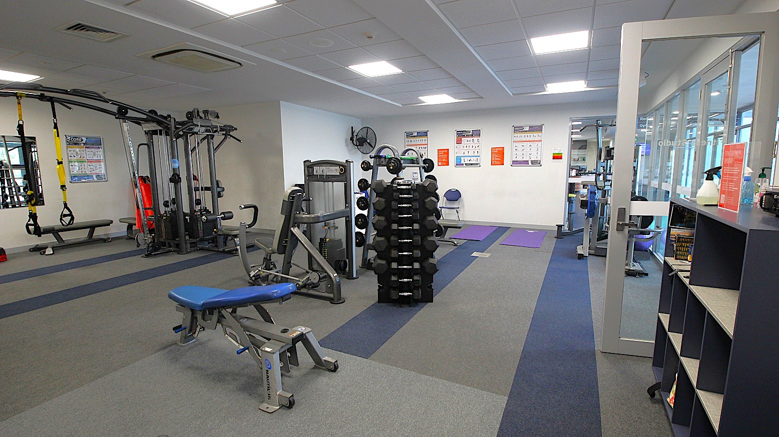 A gym with various exercise equipment. On the left, there is a multi-station machine and a bench with TRX straps hanging. In the centre, a rack of dumbbells is visible. The right side has a glass door and a shelving unit with books and cleaning supplies. The walls have instructional posters, and the floor is covered with grey and blue carpet tiles. The ceiling is fitted with fluorescent lights. A gym with various exercise equipment. On the left, there is a multi-station machine and a bench with TRX straps hanging. In the centre, a rack of dumbbells is visible. The right side has a glass door and a shelving unit with books and cleaning supplies. The walls have instructional posters, and the floor is covered with grey and blue carpet tiles. The ceiling is fitted with fluorescent lights.