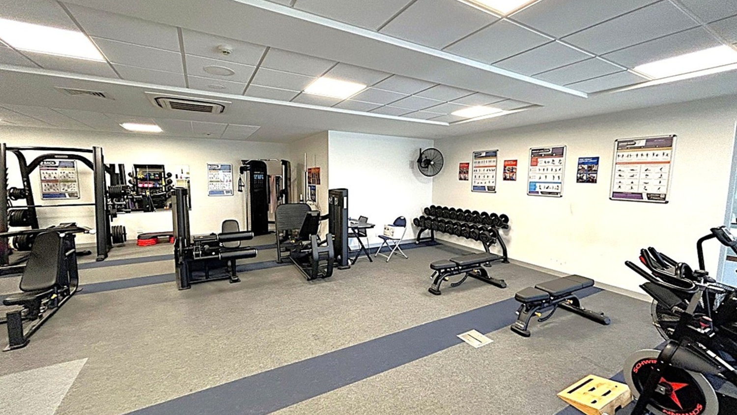 A gym with various exercise equipment. On the left, there are weight machines and a bench press. In the centre, a leg press machine and a seated bench are visible. On the right, a row of dumbbells is placed on a rack beneath wall-mounted exercise charts. A stationary bike is partially visible. The floor is carpeted in grey with a darker strip running through it. The ceiling is fitted with fluorescent lights and air conditioning vents. A wall fan is mounted near the dumbbells. A gym with various exercise equipment. On the left, there are weight machines and a bench press. In the centre, a leg press machine and a seated bench are visible. On the right, a row of dumbbells is placed on a rack beneath wall-mounted exercise charts. A stationary bike is partially visible. The floor is carpeted in grey with a darker strip running through it. The ceiling is fitted with fluorescent lights and air conditioning vents. A wall fan is mounted near the dumbbells.
