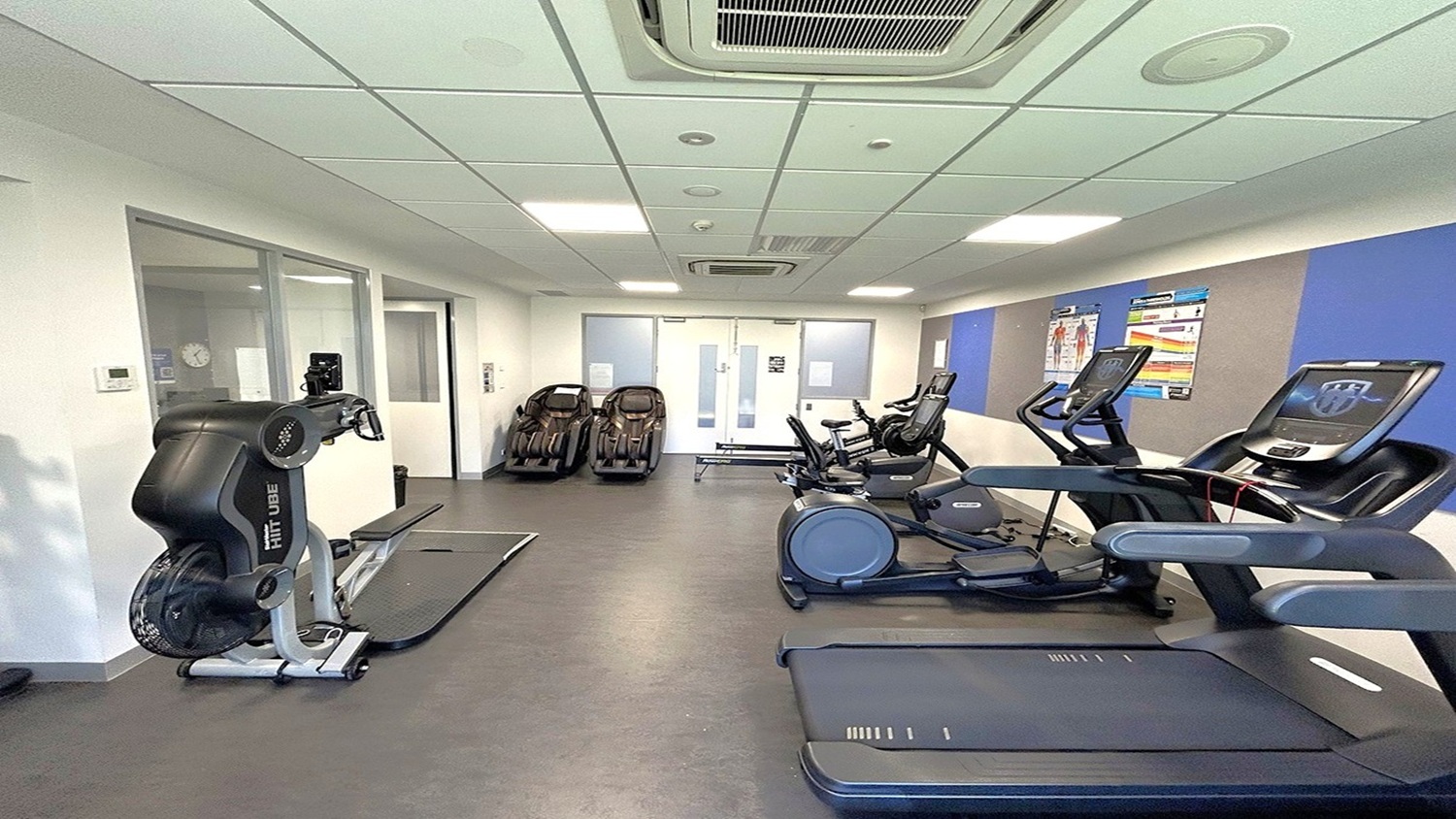 A gym room with exercise equipment. On the left, there's a FitLuge machine near a glass partition. In the middle, several cardio machines, including treadmills and exercise bikes, are lined up against the wall. On the right, two treadmills are visible. The walls have blue and beige panels with fitness posters. The floor is dark grey, and the ceiling is white with recessed lighting and air conditioning vents. A gym room with exercise equipment. On the left, there's a FitLuge machine near a glass partition. In the middle, several cardio machines, including treadmills and exercise bikes, are lined up against the wall. On the right, two treadmills are visible. The walls have blue and beige panels with fitness posters. The floor is dark grey, and the ceiling is white with recessed lighting and air conditioning vents.