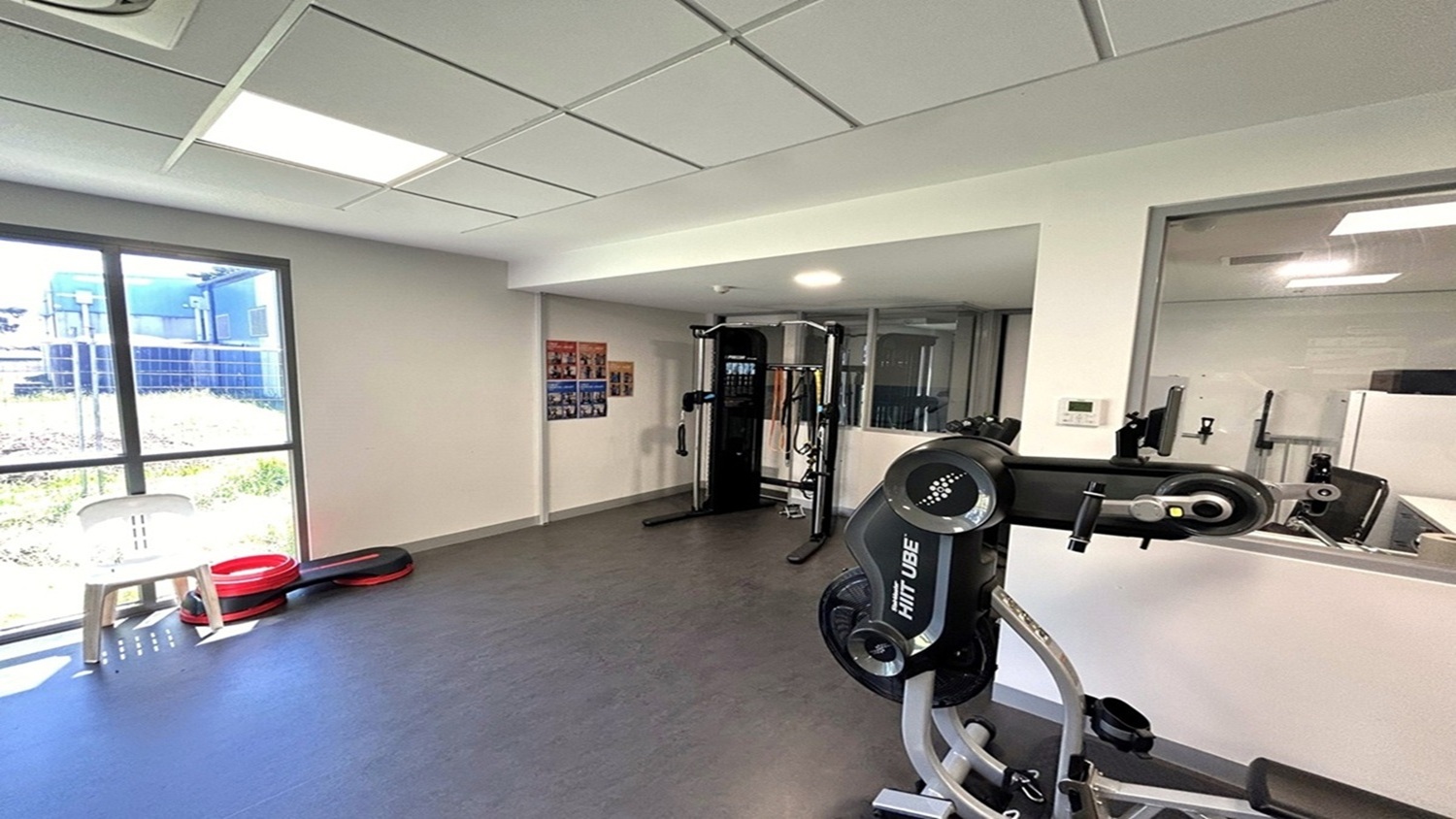 A gym room with exercise equipment. On the right, there's a large machine with "HIT UBE" written on it. In the centre, a multi-functional gym station is visible. On the left, a window allows natural light in, with a white chair and stacked red and black exercise steps nearby. The walls are light-coloured, and there are posters on the wall. The ceiling has recessed lighting, and the floor is covered with grey tiles. A gym room with exercise equipment. On the right, there's a large machine with "HIT UBE" written on it. In the centre, a multi-functional gym station is visible. On the left, a window allows natural light in, with a white chair and stacked red and black exercise steps nearby. The walls are light-coloured, and there are posters on the wall. The ceiling has recessed lighting, and the floor is covered with grey tiles.
