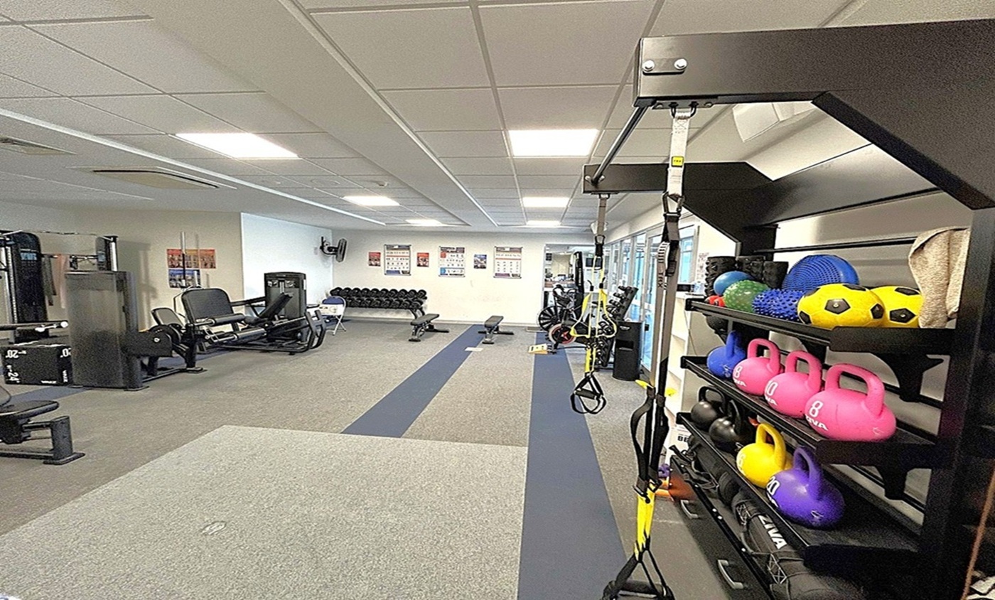 A gym interior with a variety of equipment. On the right, a rack holds colourful kettlebells, medicine balls, and other fitness gear. In the middle, a long strip of flooring leads to more equipment, including benches and weights. On the left, there are exercise machines and a row of dumbbells against the wall. The ceiling is fitted with fluorescent lights, and the floor is carpeted with different sections for various activities. Posters are visible on the walls. A gym interior with a variety of equipment. On the right, a rack holds colourful kettlebells, medicine balls, and other fitness gear. In the middle, a long strip of flooring leads to more equipment, including benches and weights. On the left, there are exercise machines and a row of dumbbells against the wall. The ceiling is fitted with fluorescent lights, and the floor is carpeted with different sections for various activities. Posters are visible on the walls.