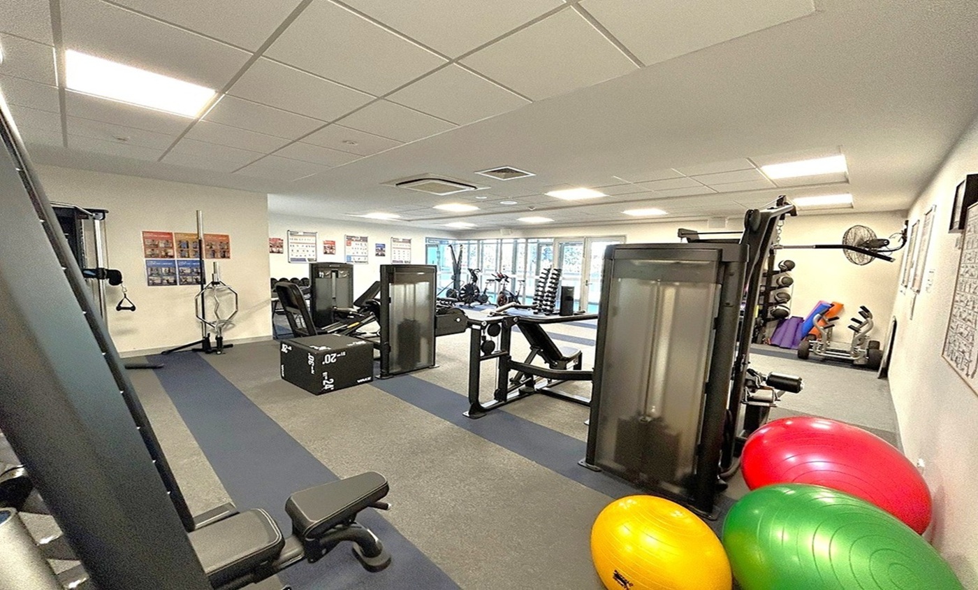 A gym with various exercise equipment. On the left, there are weight machines and a bench. In the centre, a black plyometric box is visible. On the right, there are colourful exercise balls in red, yellow, and green. The background features a wall with posters and a rack of dumbbells. The floor is covered with grey and black mats, and the ceiling has recessed lighting panels. A gym with various exercise equipment. On the left, there are weight machines and a bench. In the centre, a black plyometric box is visible. On the right, there are colourful exercise balls in red, yellow, and green. The background features a wall with posters and a rack of dumbbells. The floor is covered with grey and black mats, and the ceiling has recessed lighting panels.