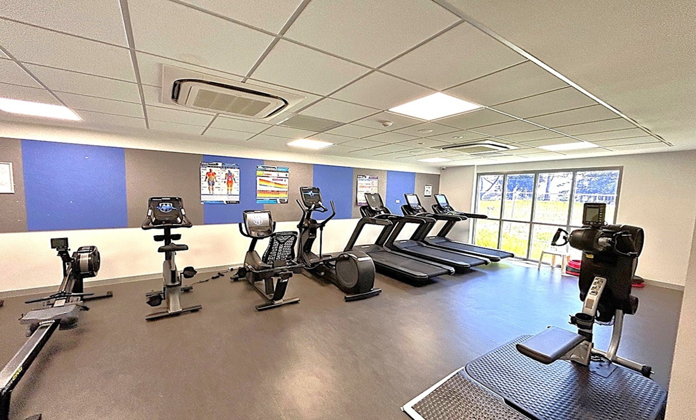 A gym with a variety of exercise equipment. On the left, there are rowing machines and stationary bikes. In the middle, several treadmills are lined up facing large windows that let in natural light. On the right, there's a piece of equipment for upper body workouts. The floor is dark and smooth, and the ceiling is fitted with white tiles and recessed lighting. The walls have blue and grey panels with fitness posters. A gym with a variety of exercise equipment. On the left, there are rowing machines and stationary bikes. In the middle, several treadmills are lined up facing large windows that let in natural light. On the right, there's a piece of equipment for upper body workouts. The floor is dark and smooth, and the ceiling is fitted with white tiles and recessed lighting. The walls have blue and grey panels with fitness posters.