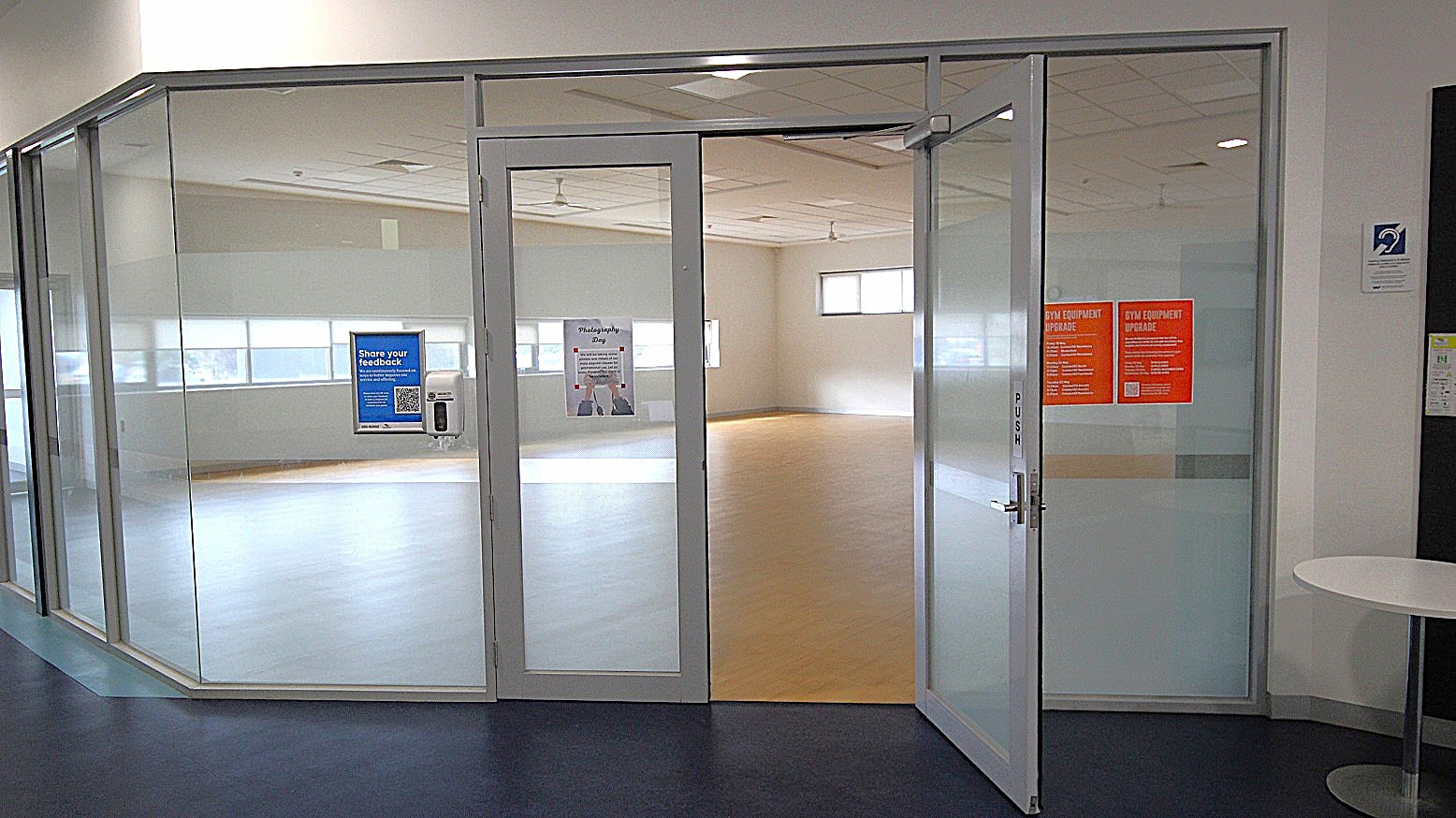 Glass-walled room with a partially open door. The door has a "PUSH" sign and displays a blue feedback poster with a QR code. Inside, the room is empty with light-coloured flooring and several windows allowing natural light. On the right, two orange notices are visible on the glass. A small round table is positioned outside the room on the right. The ceiling has recessed lighting, and the floor outside is dark. Glass-walled room with a partially open door. The door has a "PUSH" sign and displays a blue feedback poster with a QR code. Inside, the room is empty with light-coloured flooring and several windows allowing natural light. On the right, two orange notices are visible on the glass. A small round table is positioned outside the room on the right. The ceiling has recessed lighting, and the floor outside is dark.