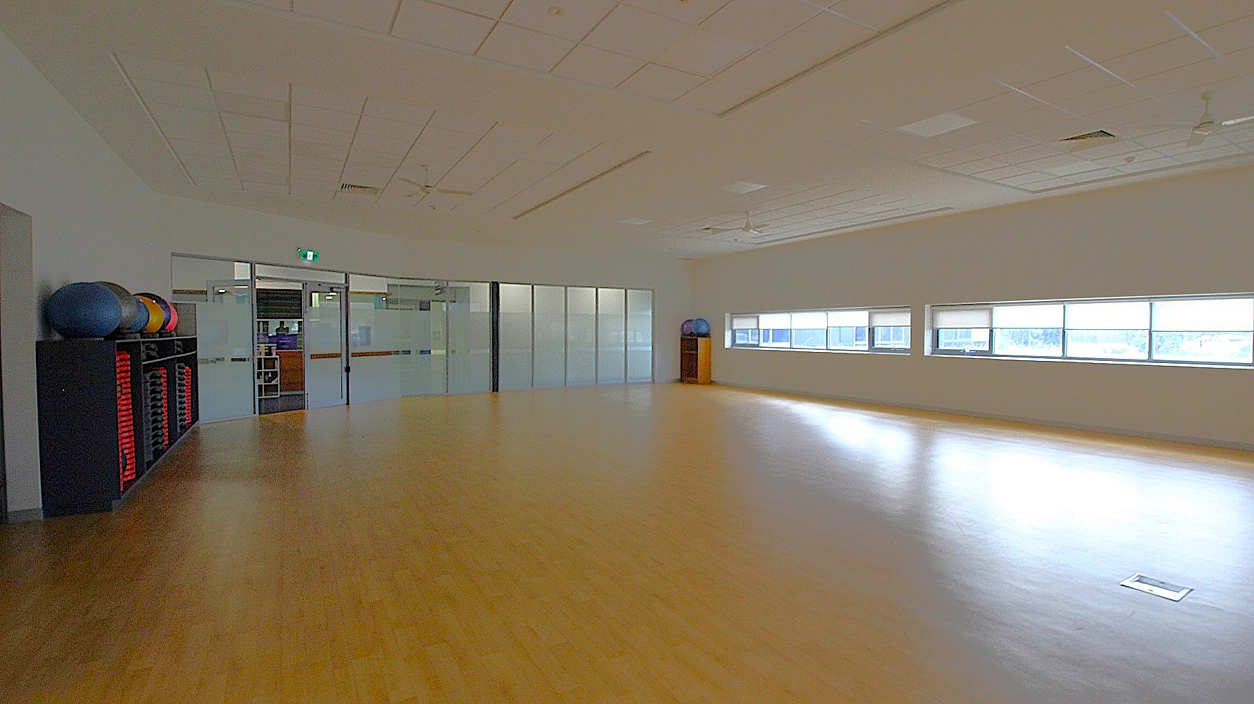 A spacious room with a light wooden floor and a white ceiling featuring recessed lighting and ceiling fans. On the left, there is a rack with exercise balls and red mats. The back wall has large glass doors and windows, allowing natural light to enter. On the right, there are more windows with a view outside. The room appears to be a fitness or dance studio. A spacious room with a light wooden floor and a white ceiling featuring recessed lighting and ceiling fans. On the left, there is a rack with exercise balls and red mats. The back wall has large glass doors and windows, allowing natural light to enter. On the right, there are more windows with a view outside. The room appears to be a fitness or dance studio.