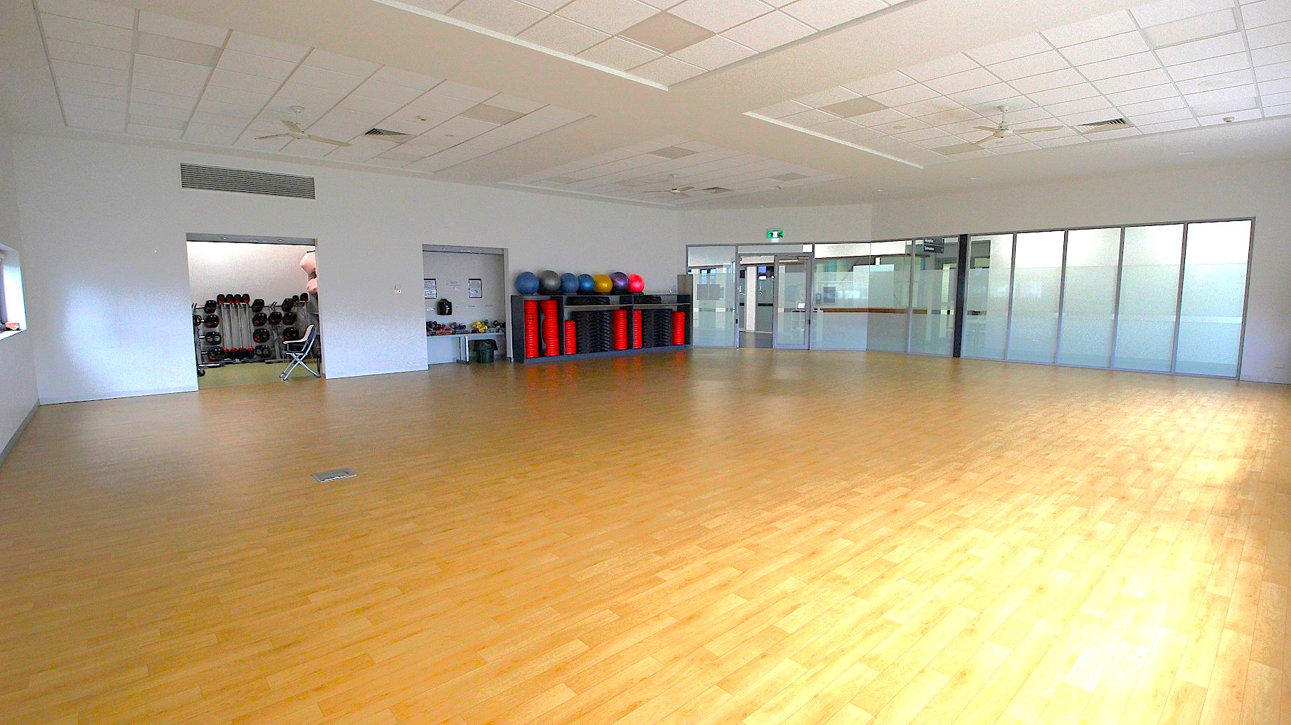 A spacious room with a light wooden floor and a white ceiling with recessed lighting. On the left, there's an open doorway leading to a storage area with exercise equipment like weights and balls. In the centre, a shelf holds various exercise balls and red mats. On the right, there are large glass doors and windows, providing a view into another area. The room is well-lit and appears to be a fitness or multipurpose space. A spacious room with a light wooden floor and a white ceiling with recessed lighting. On the left, there's an open doorway leading to a storage area with exercise equipment like weights and balls. In the centre, a shelf holds various exercise balls and red mats. On the right, there are large glass doors and windows, providing a view into another area. The room is well-lit and appears to be a fitness or multipurpose space.