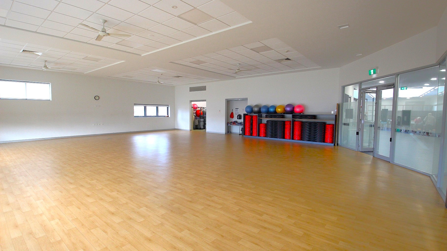 A spacious room with a light wooden floor and a white ceiling featuring ceiling fans. On the right, there are glass doors and a wall with exercise equipment, including colourful exercise balls and stacked red and black steps. The left side has two windows and a clock on the wall. The room is well-lit with natural light coming through the windows. A spacious room with a light wooden floor and a white ceiling featuring ceiling fans. On the right, there are glass doors and a wall with exercise equipment, including colourful exercise balls and stacked red and black steps. The left side has two windows and a clock on the wall. The room is well-lit with natural light coming through the windows.