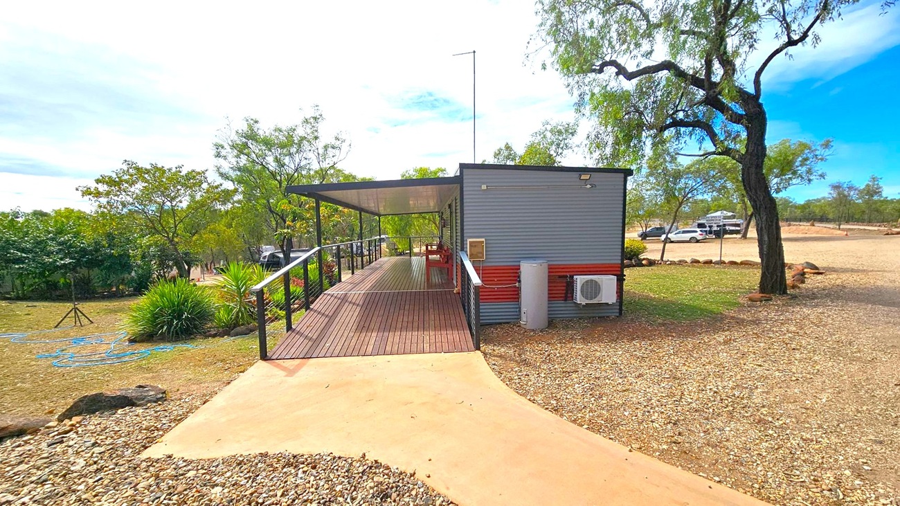 A small building with a corrugated metal exterior and a wooden deck extends from the right to the centre. The deck has a railing and a shaded area with a table and chairs. On the left, there are green shrubs and trees, with a blue hose on the ground. The foreground has a gravel path leading to the deck. In the background, a few cars are parked on a gravel area, and more trees are visible. A large tree stands on the right.