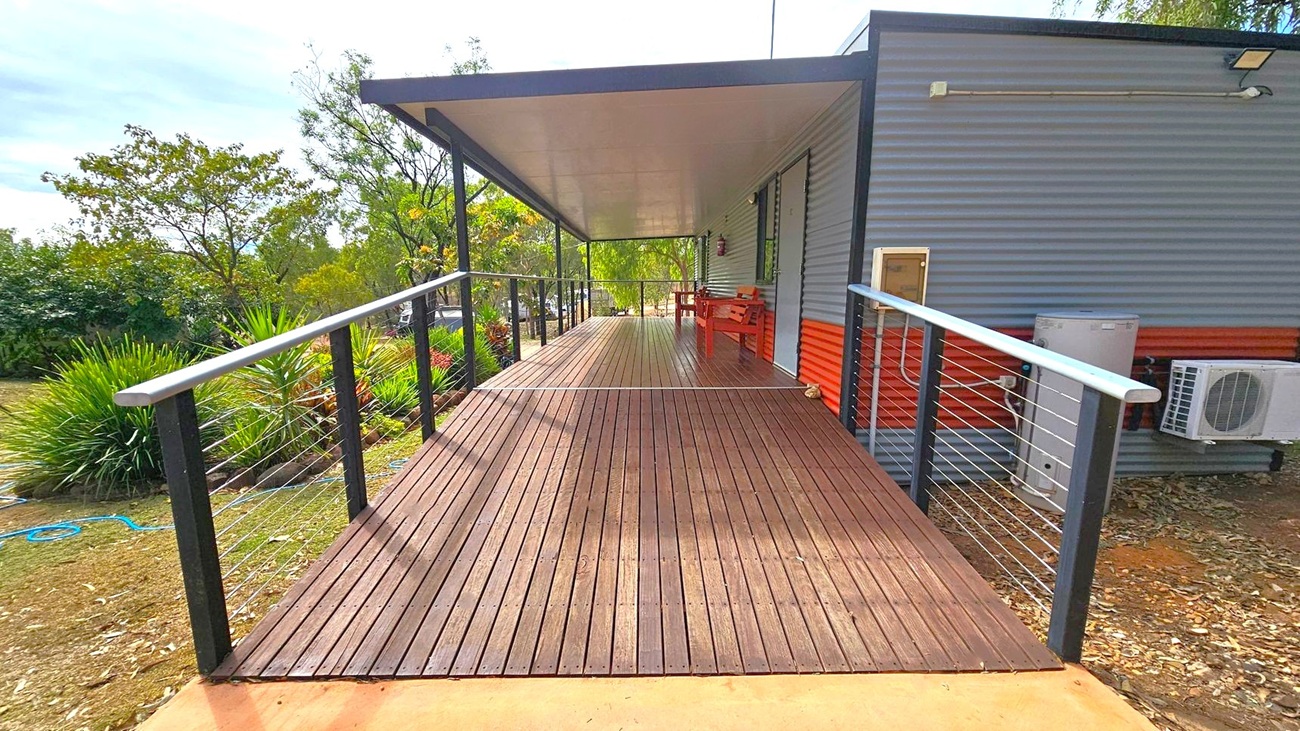 A wooden deck extends forward, bordered by metal railings with wire cables. On the right, a corrugated metal building wall features an air conditioning unit and a small bench. The deck is covered by a flat roof. On the left, there are various green plants and trees in a garden area. The ground is covered with grass and some fallen leaves.