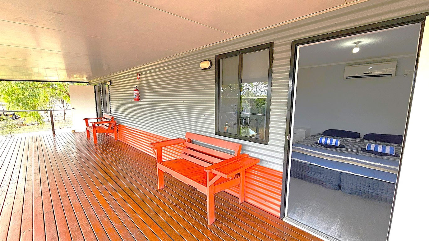 A wooden deck with a corrugated metal wall on the right, featuring a red bench and a fire extinguisher. A window and an open door reveal a room with a bed covered in striped bedding and an air conditioner above. On the left, another red bench is near the open end of the deck, which overlooks a grassy area with trees. The ceiling is smooth and light-coloured.