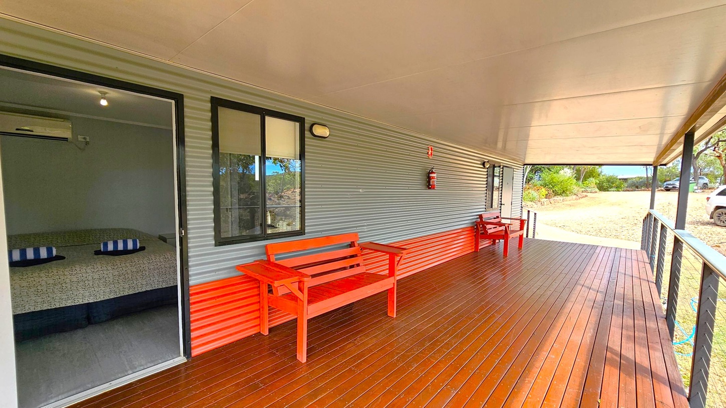 A wooden deck with a corrugated metal wall on the left, featuring a sliding glass door and a window. Inside, a bed with striped pillows is visible. Two red wooden benches are placed along the wall. The deck extends to the right, bordered by a railing. The ceiling is flat and light-coloured. In the background, there is a gravel area with parked cars and some greenery. A fire extinguisher is mounted on the wall.