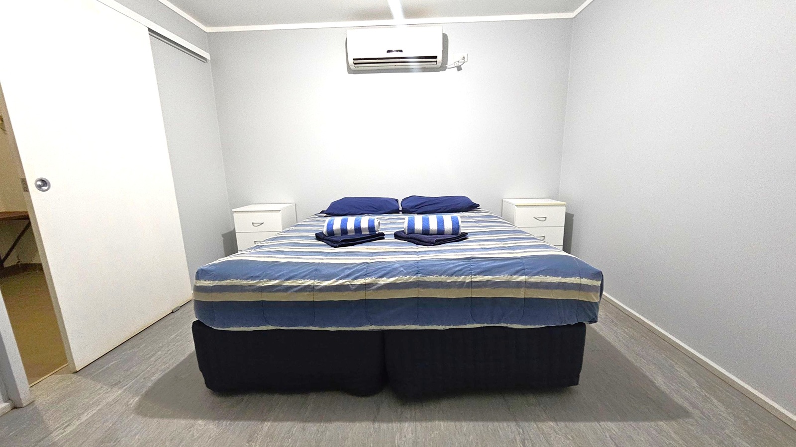 A small bedroom with a double bed in the centre, covered with a striped duvet in shades of blue and green. Two striped towels are neatly folded on the bed. On either side of the bed are white bedside tables with drawers. Above the bed is a wall-mounted air conditioning unit. The walls are plain and light-coloured. To the left, a sliding door is partially open, revealing a glimpse of another room. The floor is a light wood or laminate, and the ceiling is plain with a light fixture.