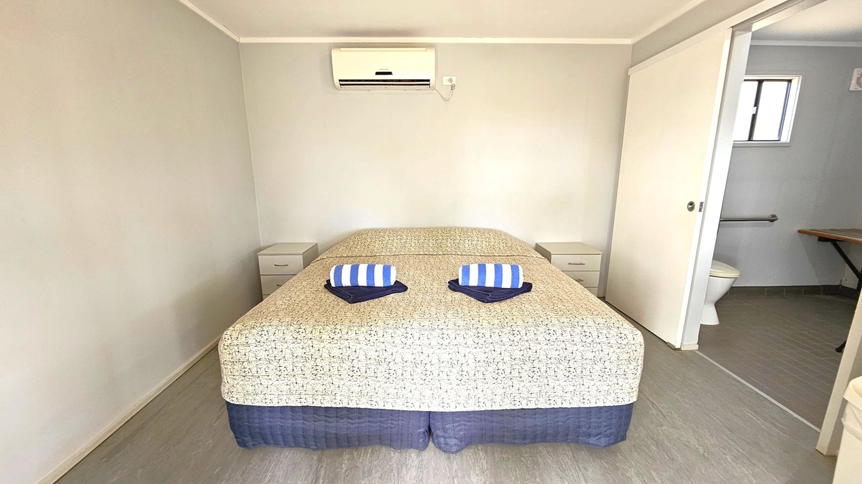 A small room with a bed in the centre, covered with a patterned bedspread and two rolled, striped towels on top. On either side of the bed are small bedside tables with drawers. Above the bed is a wall-mounted air conditioning unit. To the right, a sliding door is partially open, revealing a bathroom with a toilet and a window. The floor is covered with light-coloured tiles, and the walls are painted in a pale shade.
