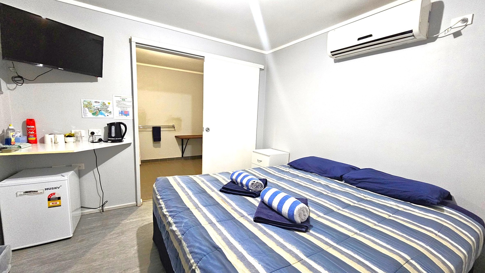 A small room with a striped bed in the foreground, topped with two rolled towels. On the left, a wall-mounted TV is above a shelf holding a kettle, cups, and condiments. Below the shelf is a mini fridge. A sliding door in the centre leads to another room with a towel on a rail. An air conditioning unit is mounted on the right wall. The floor is light-coloured, and the ceiling is plain with a light fixture.