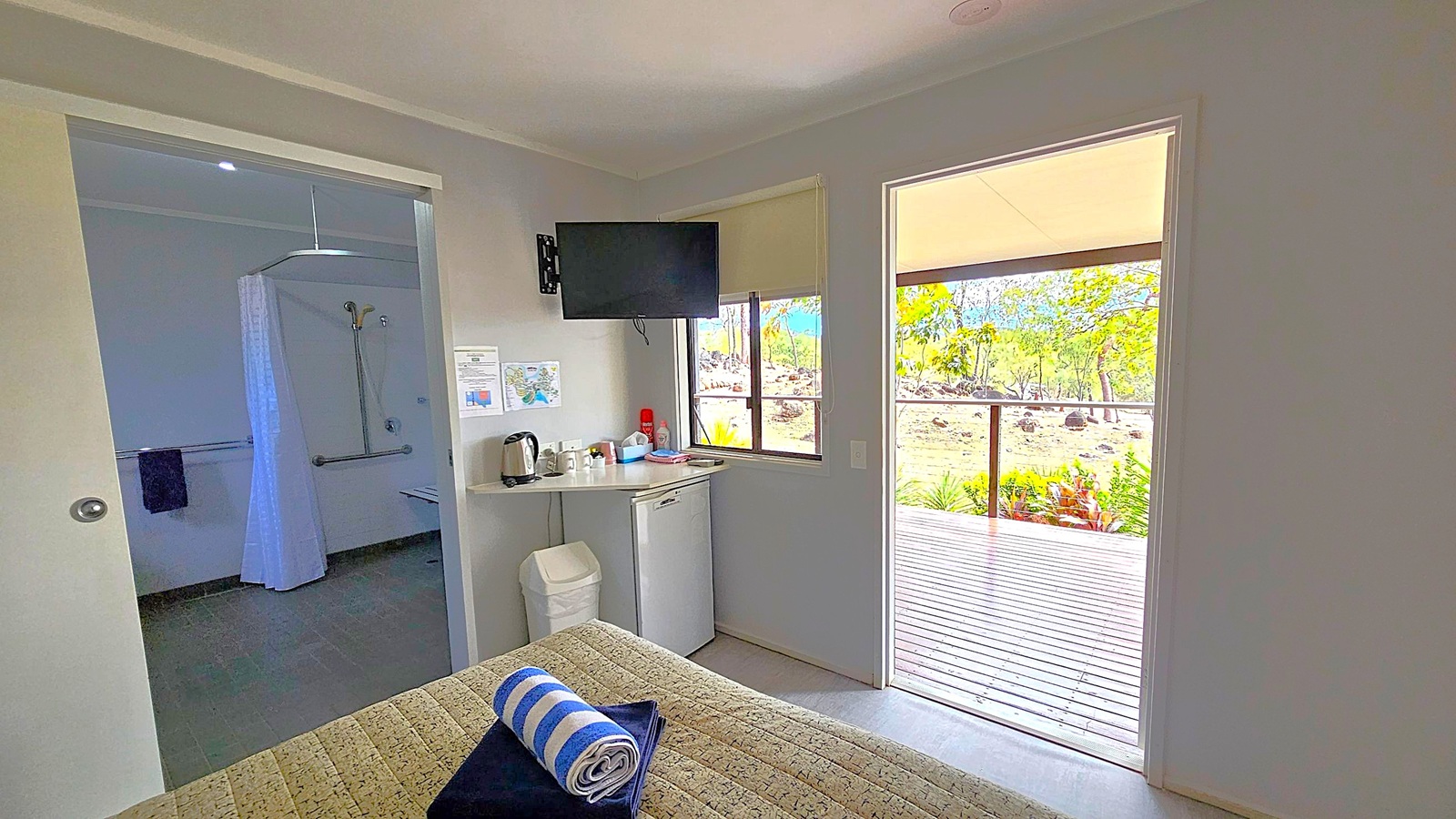A room with a view of a wooden deck and greenery outside. On the left, there's an open door leading to a bathroom with a shower and curtain. In the centre, a small counter holds a kettle and cups, with a mini fridge below. A wall-mounted TV is above the counter. A bed with a patterned cover and a rolled towel is in the foreground. The walls are light-coloured, and the ceiling is plain.