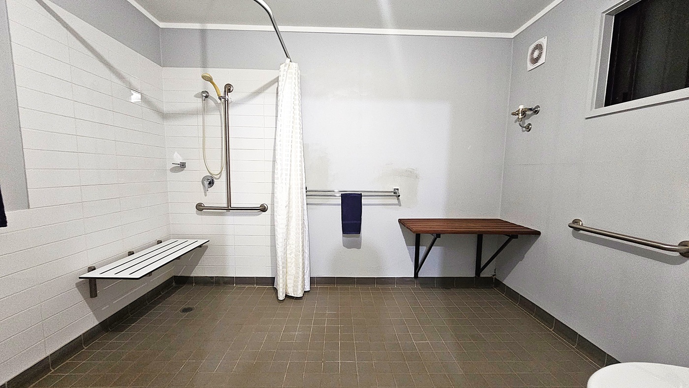 A bathroom with a green tiled floor and white walls. On the left, there is a shower area with a white curtain, a handheld showerhead, and a foldable bench. A grab bar is mounted on the wall. In the centre, a towel rail holds a dark towel. On the right, a wooden shelf is attached to the wall, and a small window is above it. A grab bar is also visible on the right wall. The ceiling is plain white.